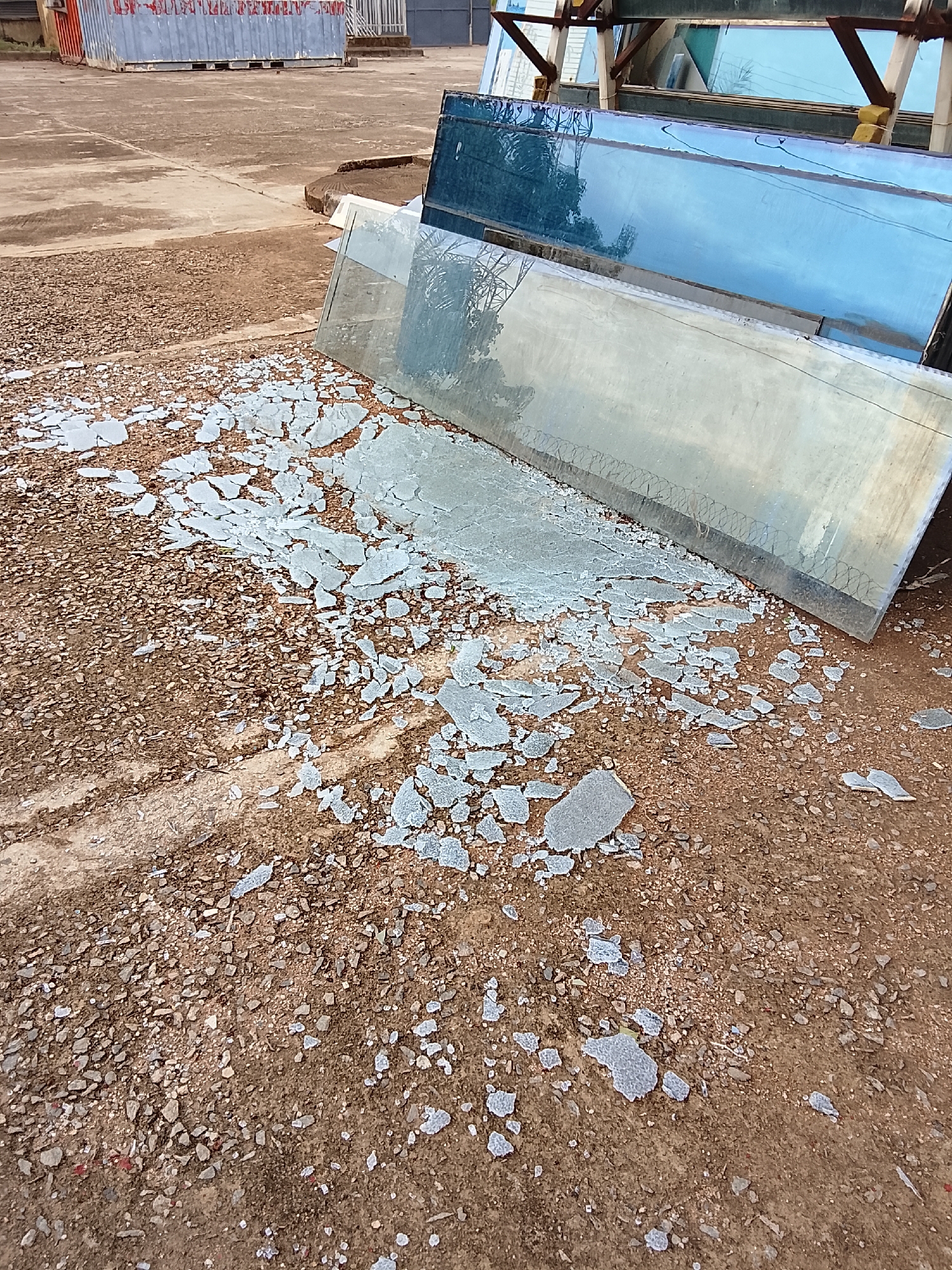 The image shows a brown, dirt ground covered with gravel and shattered glass. The glass shards are concentrated near the edges of several large panes of stacked glass, which are leaning against a metal support structure. The background includes a concrete surface and a blue cargo container. The location is most likely a storage or industrial area in Abuja, Nigeria, given the image's origin. No people are visible.