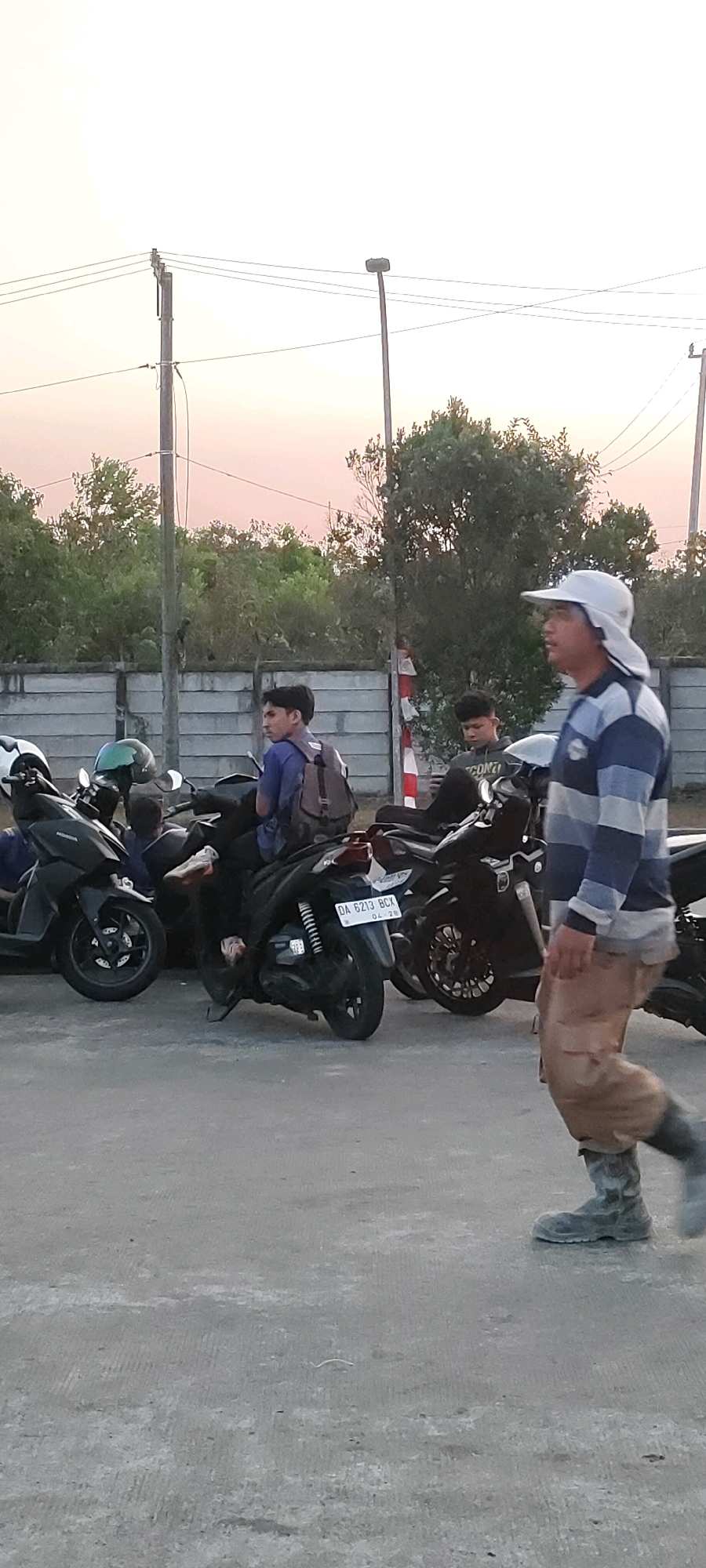 The image depicts a street scene in Bentok Kampung, Indonesia, possibly during dusk, with a warm, hazy sky in the background. In the foreground, a man wearing a light-colored wide-brimmed hat, a blue and white striped long-sleeved shirt, and khaki-colored pants with boots is walking from right to left. He appears to be a construction worker or someone in a similar profession, as indicated by his attire. Behind him, a row of motorcycles is parked. One motorcycle in the middle ground has a license plate that reads "DA 6213 BCX." A young man with a backpack is seated on this motorcycle. To the left of this motorcycle, another young man wearing a blue shirt and a backpack is visible. Further to the left, parts of other motorcycles and helmets are visible. The background features a concrete wall, followed by a dense line of trees and vegetation. Utility poles and overhead wires are also present in the upper portion of the image. The ground is paved with concrete.