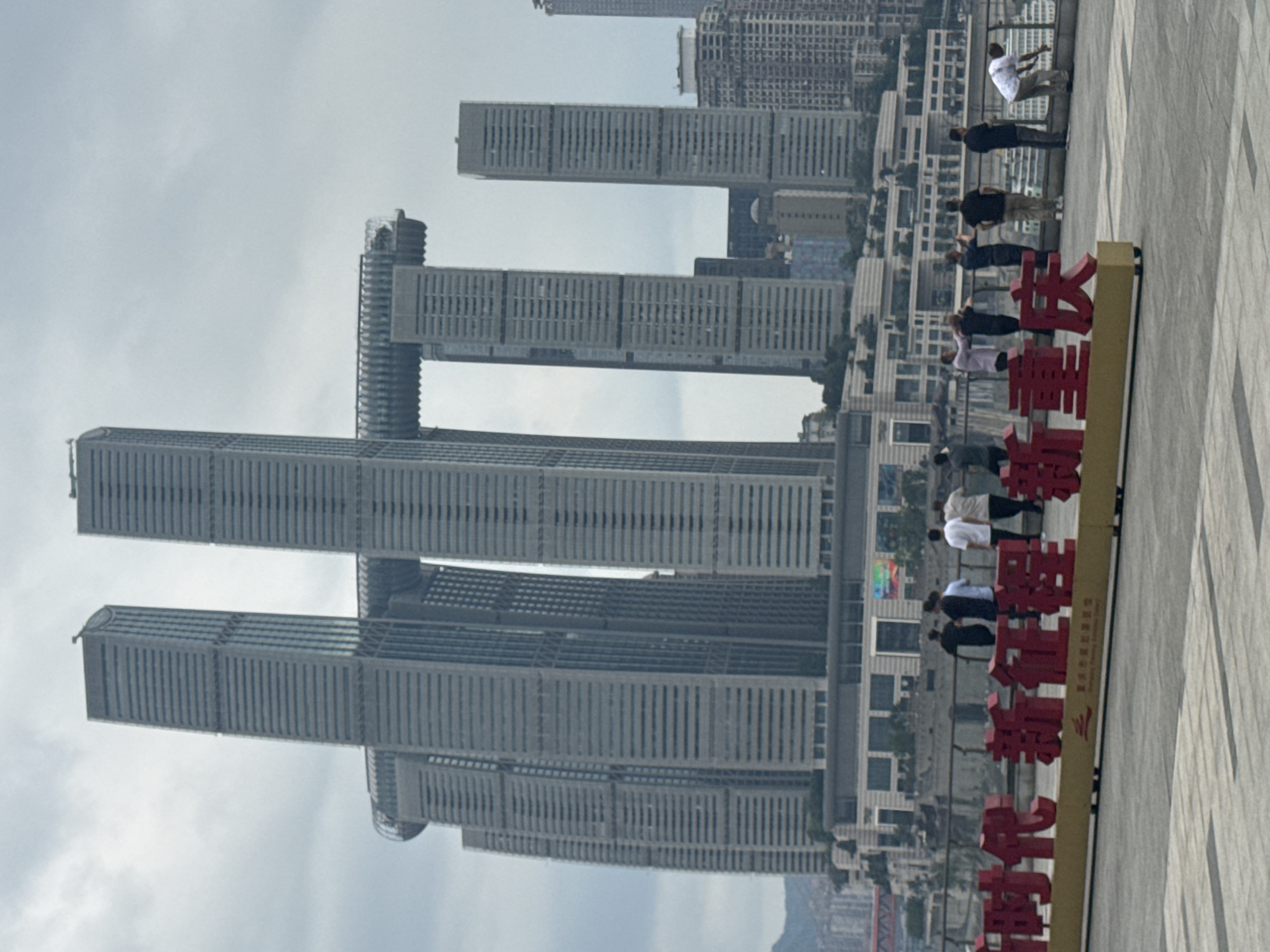 The image shows a view of the Raffles City Chongqing complex in Nan'an District, China. The complex consists of eight skyscrapers, with four taller towers and four shorter towers connected by a sky bridge. The photograph is taken from an elevated position looking down at the complex. A group of people stands on a pedestrian walkway next to a large sign with Chinese characters. The background includes other buildings and city infrastructure. The sky is overcast.
