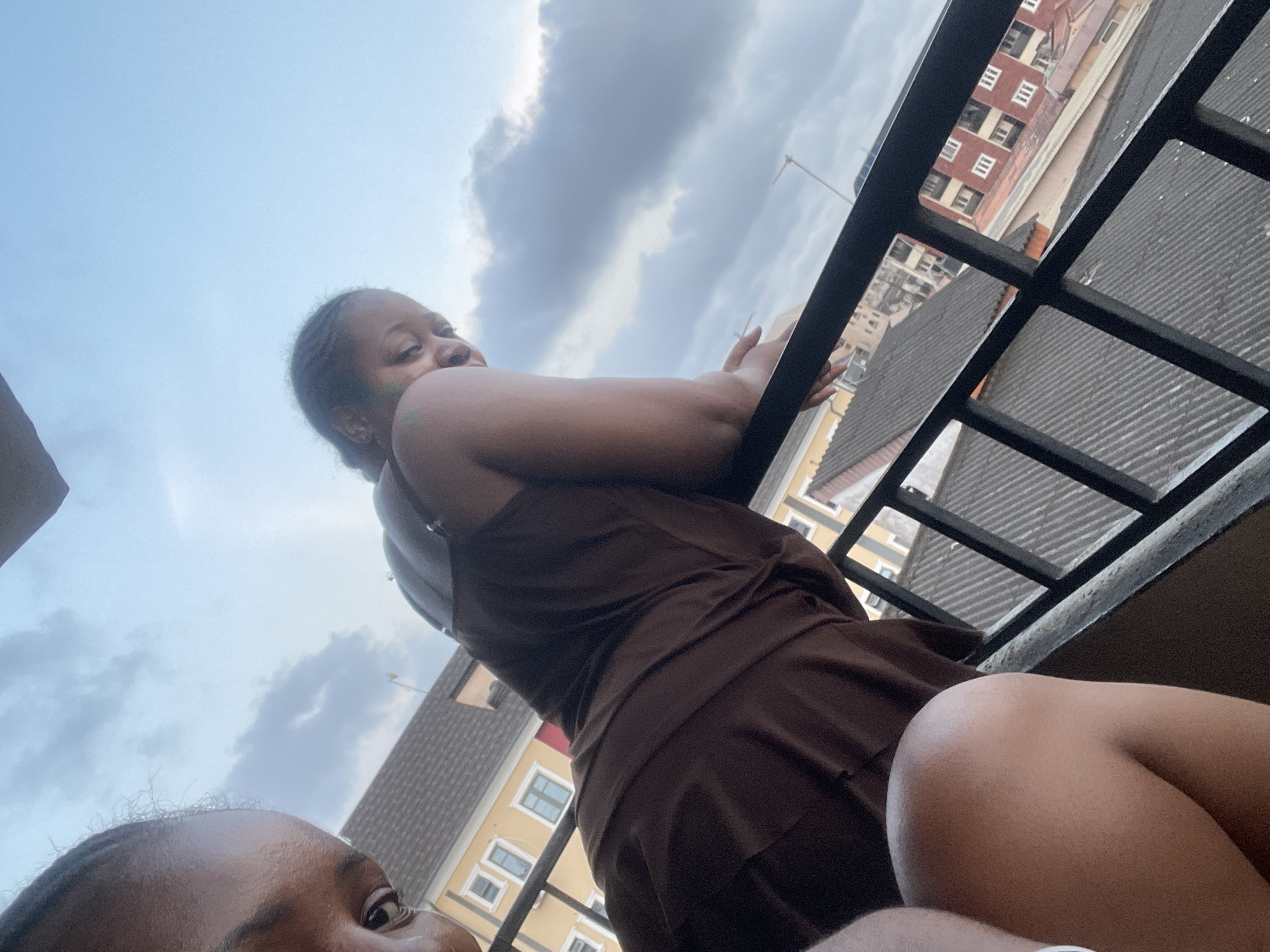 The image depicts two women on a balcony overlooking a cityscape. The woman in the foreground, on the left, is partially visible, with her eye and forehead in focus. The second woman is in the mid-ground, leaning on a black metal railing, looking away from the camera. She is wearing a brown sleeveless dress. Behind them, buildings with red and yellow facades are visible, along with corrugated metal rooftops, characteristic of the urban landscape of Animashawun, Nigeria. The sky is overcast with grey clouds.