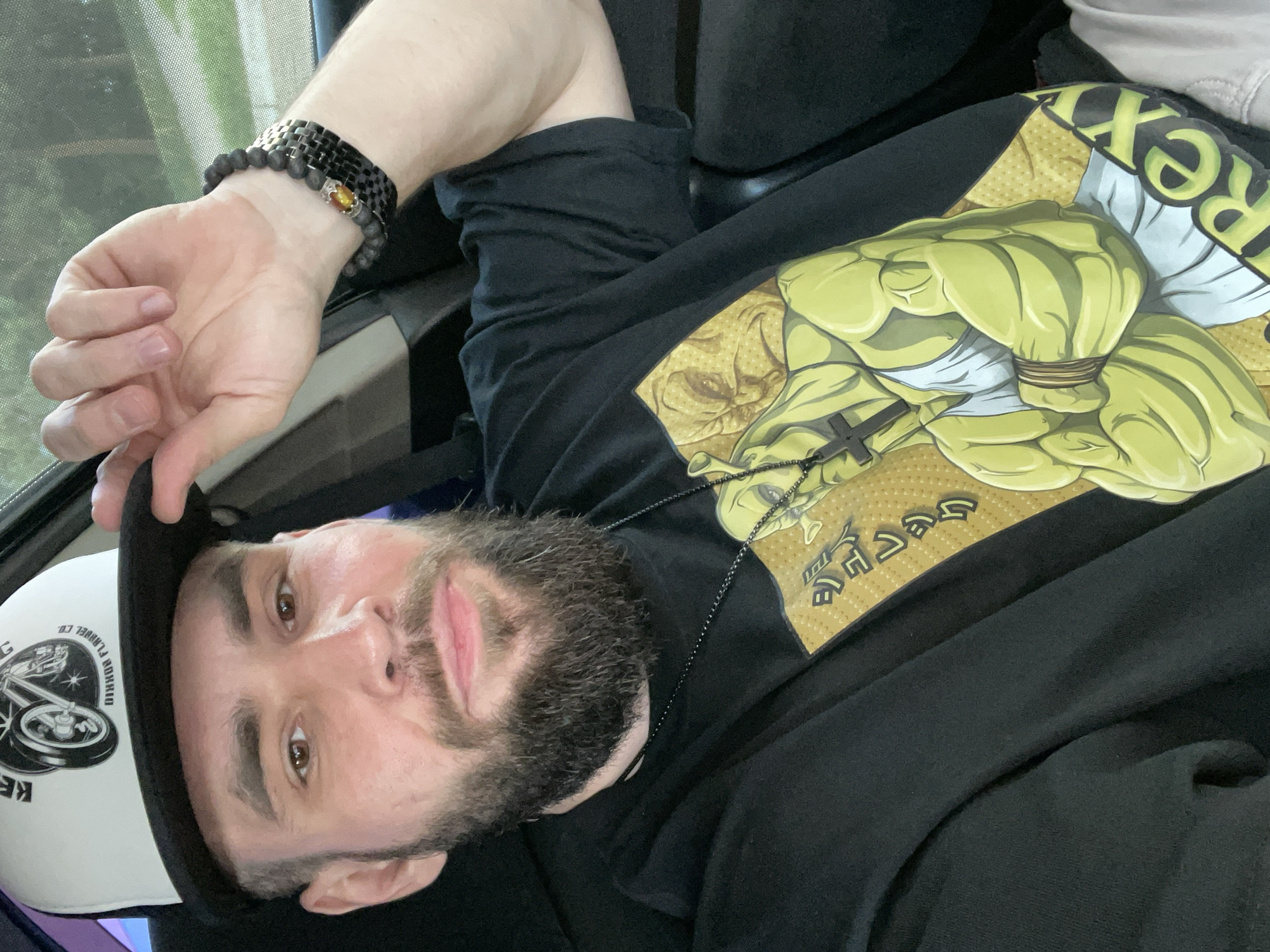 The image shows a close-up, vertically oriented selfie of a man with a beard and brown eyes. He is wearing a black t-shirt with a graphic design of the Hulk's muscular arms. The Hulk's arms are depicted in a lime green color, with white bands around the wrists. A small black cross pendant hangs from a chain around his neck, resting on the t-shirt. The man is also wearing a black baseball cap with a white front panel. On the front panel of the cap, there is a circular logo with a wheel and text that appears to be in English. He is holding the brim of the cap with his left hand, which has two bracelets on the wrist. One bracelet is black beaded, and the other appears to be a linked metallic band with a dark stone. The man's expression is neutral, with his mouth slightly open. The background is blurred but suggests an indoor setting, possibly a car or bus, with hints of greenery visible through a window. The lighting suggests it is daytime. No specific time of day or weather conditions are discernible. The text visible on the cap reads "KEHRIN FLUNNEL CO." and some other unreadable words in a circular pattern.