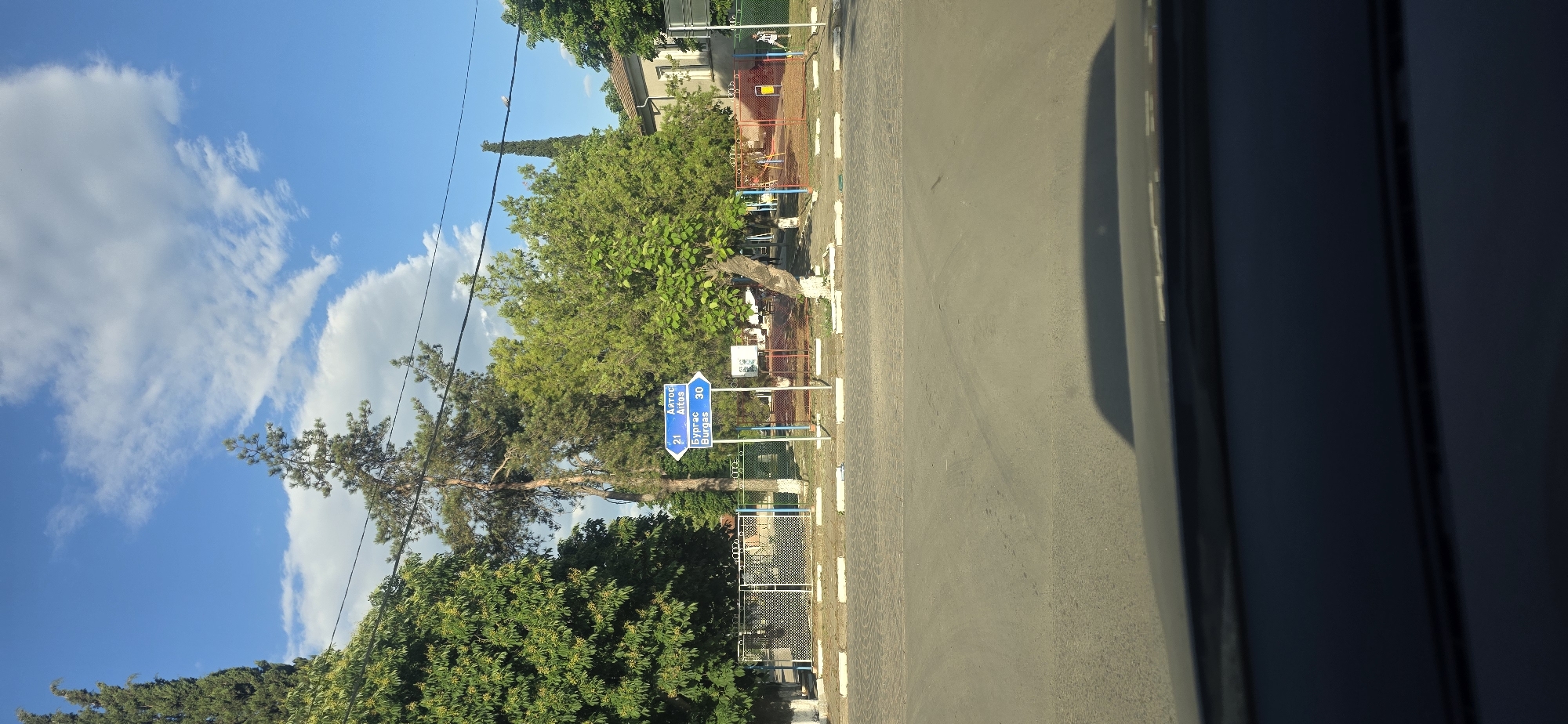 The image shows a roadside scene in Troyanovo, Bulgaria. A blue directional sign indicates that Aytos is 21 km to the left and Burgas is 30 km to the right. The sign is positioned near a fenced area with trees and possibly a playground. The sky is blue with clouds. The view is taken from inside a vehicle, indicated by the partially visible car door frame. The road occupies the right side of the image.