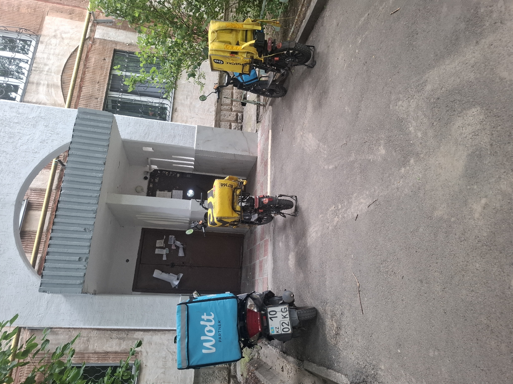 The image depicts an apartment building entrance in Almaty, Kazakhstan, with three parked delivery scooters. Two scooters are positioned in front of the building's doors, and one is to the right. The scooter on the left is blue and white and displays the "Wolt Partner" logo. The scooter in the center is yellow and has "Yandex.Eda" branding visible. The scooter on the right is also yellow with "Yandex" branding. The building has a textured white facade with a grey corrugated metal overhang. The ground is paved with asphalt. Green foliage is visible to the left and right of the building entrance. The scene appears to be an outdoor, urban environment.