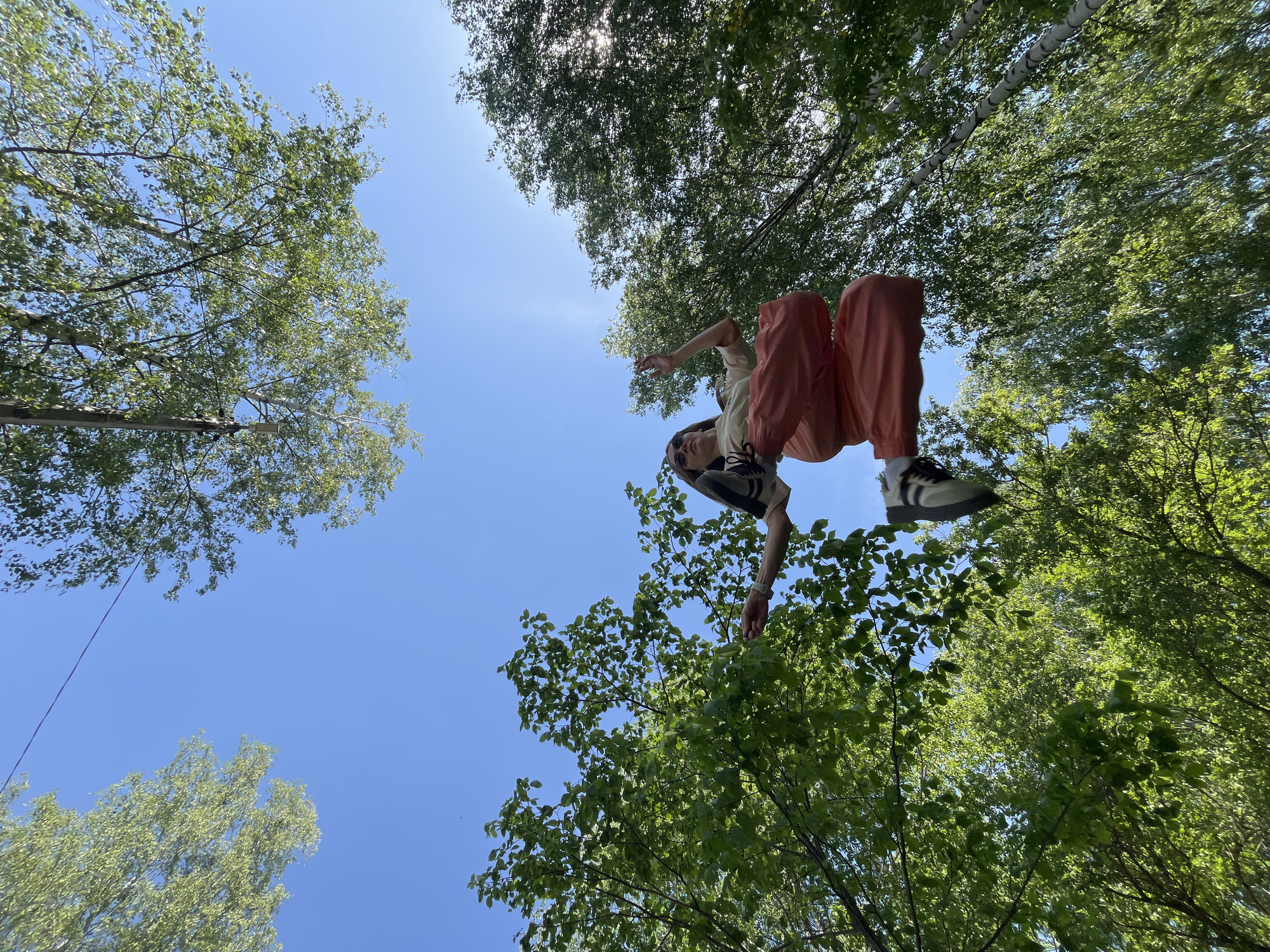 A low-angle, wide shot depicts a person suspended in the air within a forested area. The individual wears a light-colored shirt, orange pants, and black and white shoes. The person's head is oriented downwards, with one arm extended upwards and the other downwards. The background is filled with trees and a clear blue sky. A rope can be seen on the left side of the frame, extending towards the sky. The photograph was taken in the city of Chaikovsky, Russia.