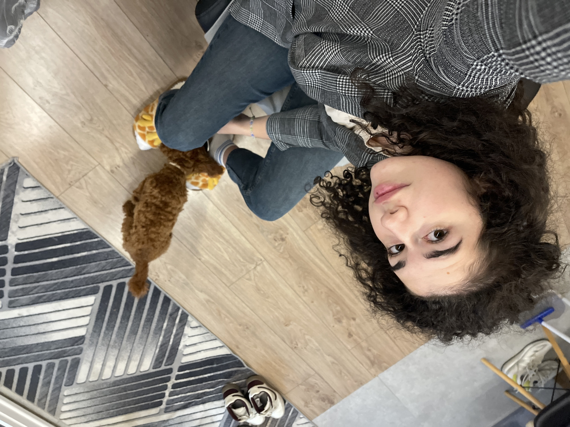 This overhead shot, taken in Sofia, Bulgaria, captures a young woman and a small dog in a casual indoor setting. The woman is seated on a light wooden floor, her gaze directed upwards towards the camera with a neutral to slightly contemplative expression. She has prominent dark, curly hair and is dressed in a grey and white plaid shirt, dark blue jeans, and light-colored socks. On her feet are distinctive yellow and brown animal-themed slippers.
A small, light brown dog with curly fur is positioned near her feet, appearing to interact with or sniff one of the slippers. The flooring transitions from the light wood in the foreground to a grey tiled area in the background. A modern rug featuring a geometric pattern in shades of grey, black, and white partially covers the wooden floor. In the wider shot, glimpses of white sneakers, a chair leg, and a cleaning tool with a blue and white handle are visible in the background. The scene suggests a relaxed, daytime moment at home. There is no visible text in the image.