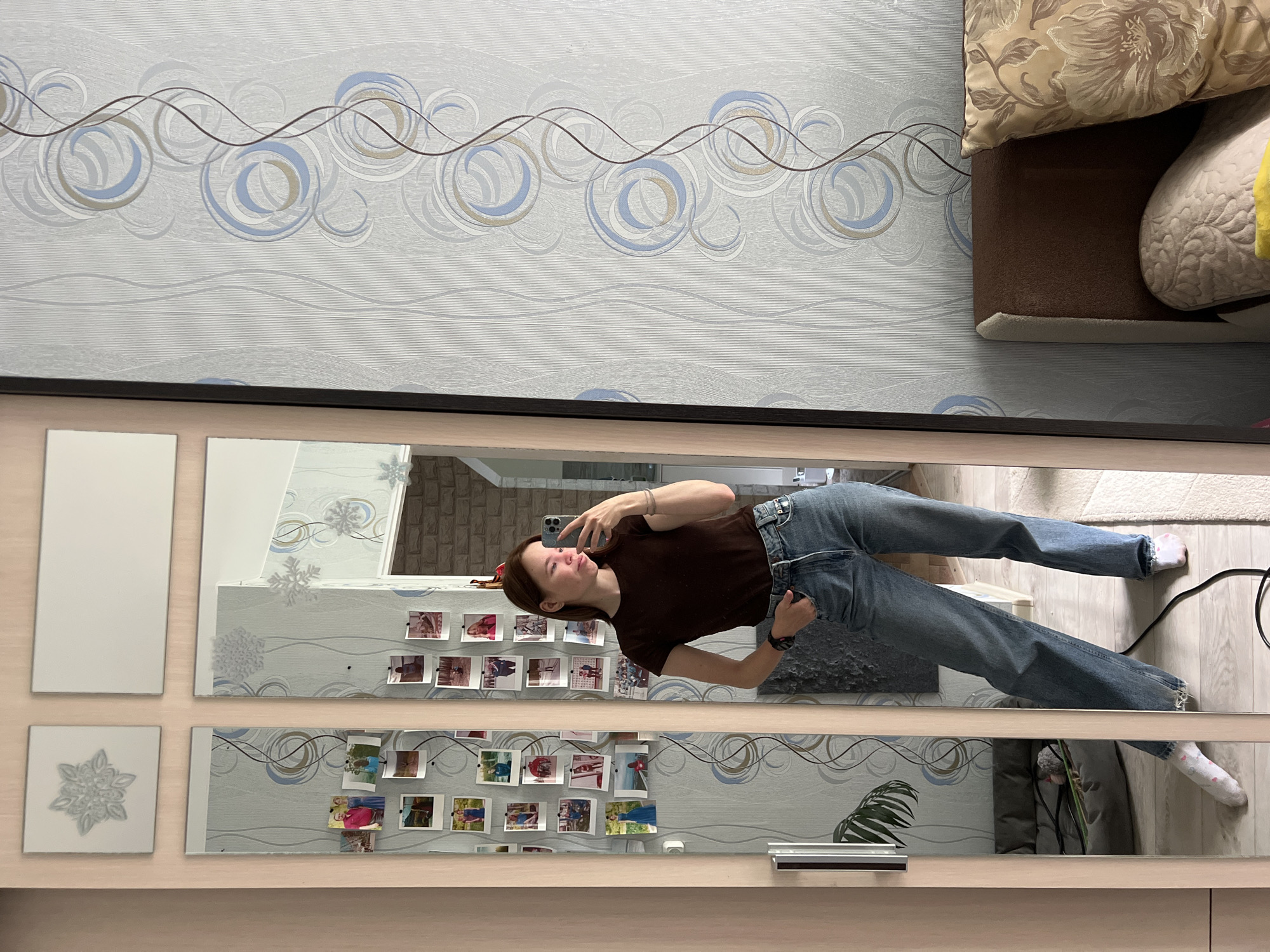 An interior shot, rotated 90 degrees counter-clockwise, depicts a young woman taking a mirror selfie in what appears to be a bedroom or living area in Chaikovsky, Russia.
The central subject is a woman with dark brown hair, wearing a dark brown t-shirt and light blue jeans, holding a light-colored smartphone to take her picture. She is reflected in a large, multi-paneled mirror that forms a significant part of the room's decor. She wears light-colored socks, one of which has pink dots, and a bracelet on her left wrist. Her expression is calm, with a slight smile.
The mirror's surface is adorned with numerous small, square printed photographs, possibly instant prints, attached in rows. These photos depict various people and scenes, suggesting personal memories. Decorative white snowflake cutouts are also affixed to some mirror panels. To the left of the main mirror, two blank rectangular white panels are visible, one featuring a raised snowflake design.
The room's walls are covered in light grey wallpaper with a distinctive pattern of swirling circles in shades of blue, grey, and beige, interwoven with wavy brown lines. In the background, reflected in the mirror and partially visible directly, is a brown couch or bed with a floral-patterned beige cushion and a textured white pillow. The floor appears to be a light wood or laminate, with a white rug near the woman's feet. A dark brown frame borders the mirror panels. A green plant, possibly a palm frond, is visible as a reflection in the lower right portion of the mirror. The overall setting suggests a well-maintained and personalized domestic space. The lighting appears bright and uniform, indicating daytime or adequate artificial illumination. No prominent visible text is discernible in the image.