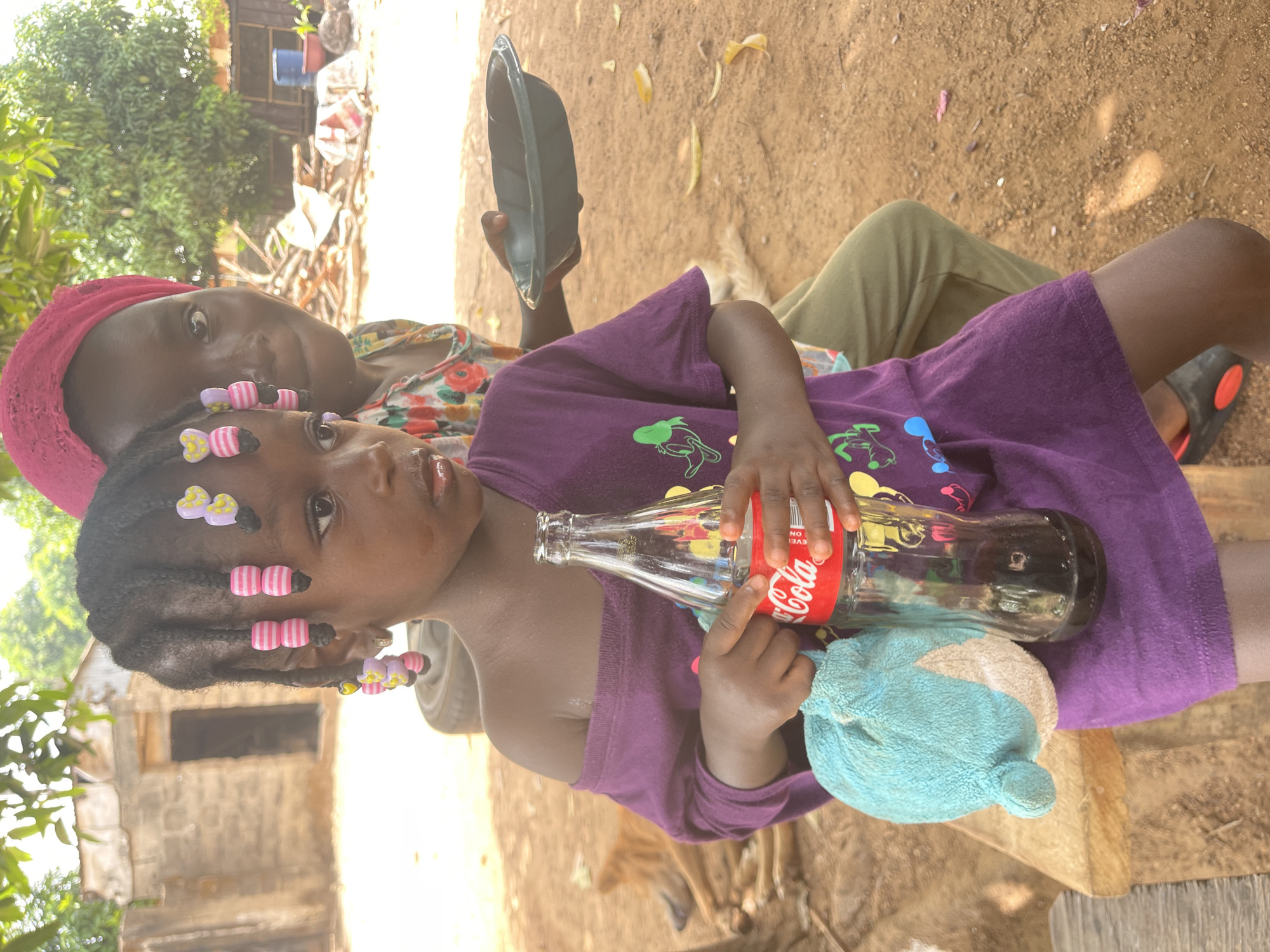 A young child with braids adorned with beads and wearing a purple shirt is being held by an adult wearing a pink headscarf. The child is holding a glass bottle of Coca-Cola and a blue plush toy. In the background, another adult is holding a black plate. The setting appears to be outdoors in a rural area, possibly in or near Akwanga, Nigeria, given the presence of basic brick structures and the overall environmental conditions. The scene is captured during daylight hours.