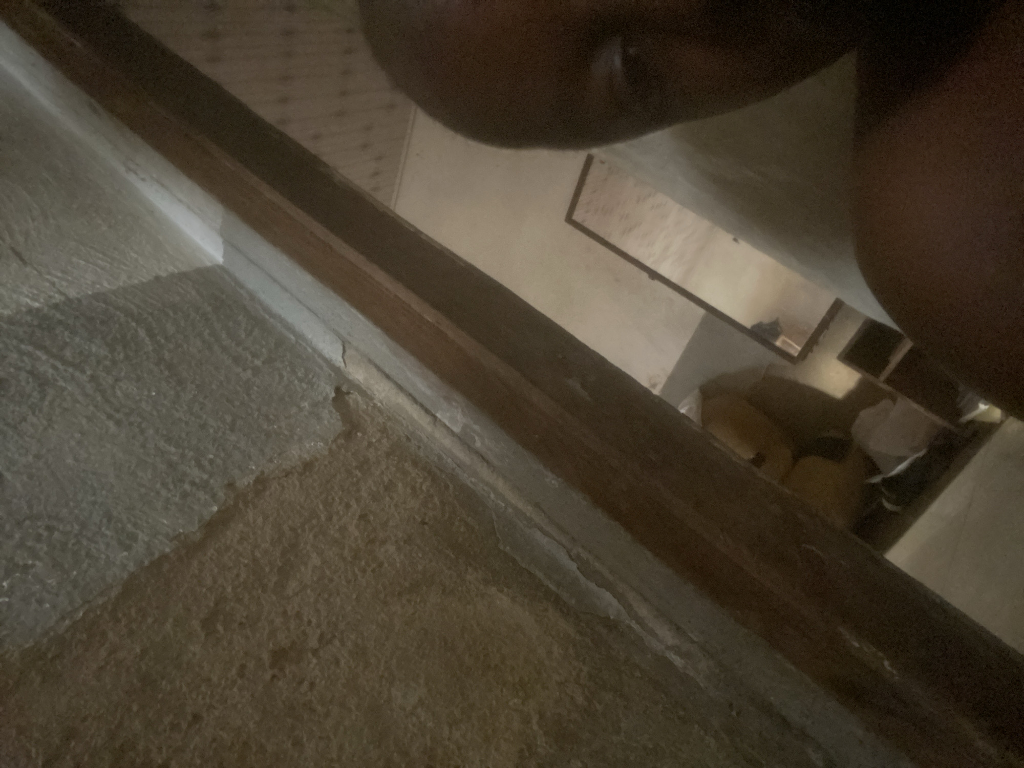 The image depicts an indoor scene, possibly a room in Jauro Yinu, Nigeria. The foreground is dominated by a textured, off-white floor and a wooden frame running diagonally across the image. Beyond the wooden frame, a tiled floor is visible, interspersed with darker rectangular sections. In the mid-ground, a mirror reflects a dimly lit area containing what appears to be furniture or stored items. The upper portion of the image is blurred and shows a dark, organic shape, possibly a person's body part, suggesting a close-up perspective. The lighting is low, creating shadows and obscuring finer details. The overall impression is of a somewhat unkempt or partially furnished space.