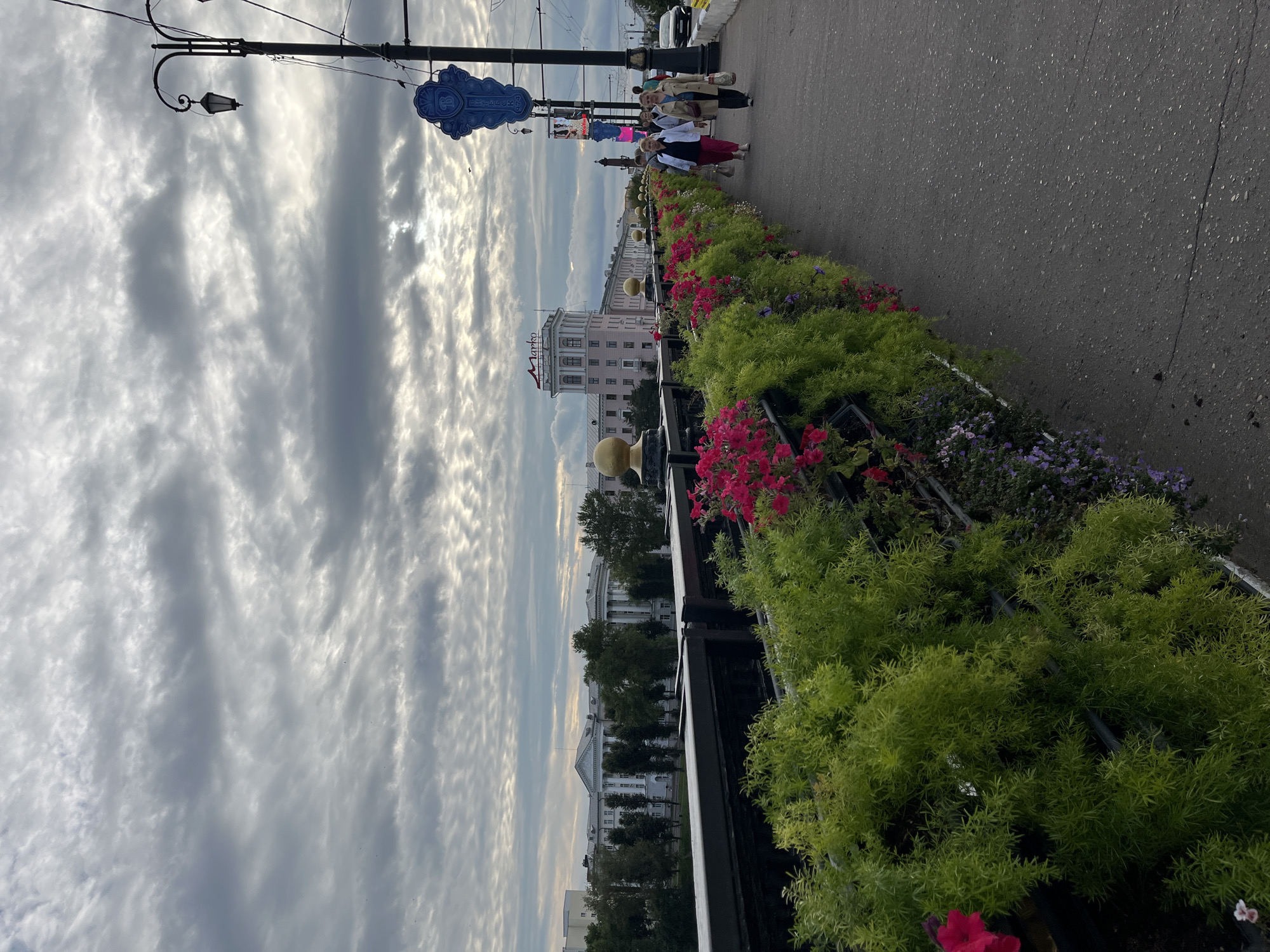 A view from a bridge in Vitsebsk, Belarus, looking towards a street with buildings and a cloudy sky. On the bridge, there is a pedestrian walkway lined with greenery and flowers. Several people are walking on the path. A lamppost with a decorative element is visible on the right side of the image. In the background, buildings of various architectural styles are seen, along with trees. The sky is filled with textured, layered clouds.