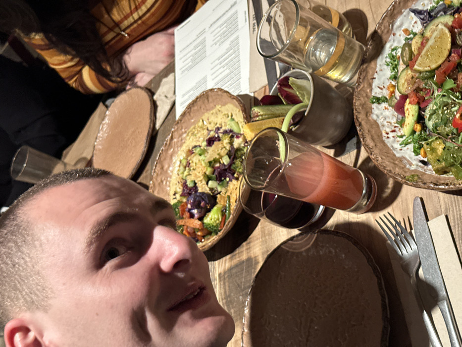 A person with short hair is taking a selfie at a restaurant. The table displays two plates of food, including a salad with avocado and a dish with couscous and vegetables. There are also several glasses, one filled with a red liquid, another with a clear liquid, and a small container with vegetables. Cutlery sits to the right of the dishes. A portion of another person is visible at the top of the frame.