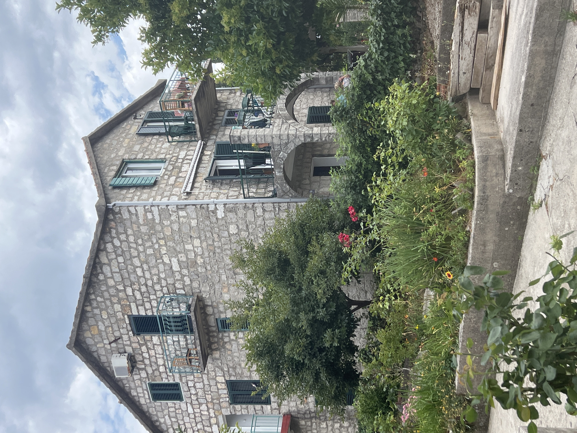 An outdoor, eye-level shot captures a multi-story stone building with a lush garden and cloudy sky, located in Baošići, Montenegro. The building features a traditional stone facade with varied stone sizes, a gabled roof, and numerous windows fitted with green louvered shutters. Several small balconies with metal railings extend from the facade. On the right side, two prominent arched openings are visible, one at ground level and another on an upper floor. An air conditioning unit is mounted on the left side of the building.
The foreground and right side are dominated by a vibrant garden with dense green foliage, including several trees and various bushes. Red and yellow flowers are scattered among the greenery. Concrete steps and a low concrete wall ascend along the right edge of the frame, with some wooden planks stacked nearby.
Visible domestic activity includes laundry hanging on a balcony railing on the left side of the building. On the upper right balcony, partially obscured by greenery, two individuals or figures in light-colored clothing are present. Another individual in a light blue top is visible through the lower arched opening on the right side. The sky is overcast with visible clouds, indicating a bright but cloudy day. No text is visible in the image.
