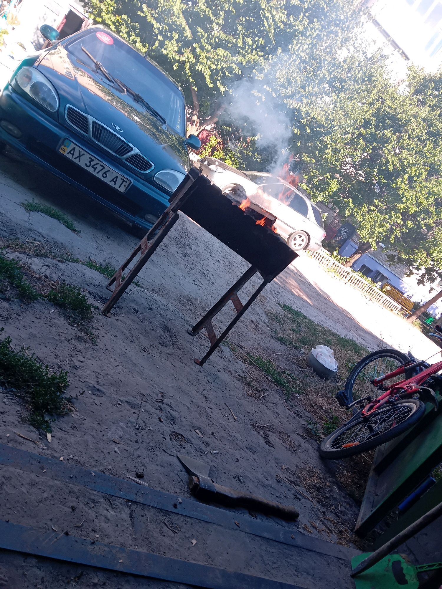 This image captures an outdoor scene on a sunny day in the city of Chuhuiv, Ukraine. In the foreground, a rusty metal barbecue grill stands on a dirt and gravel surface. Smoke rises from the grill, indicating a fire is in progress. To the left, the front of a dark teal car with the license plate "AX 3476 AT" is visible. Behind the grill, a light-colored car is parked. Further back, lush green trees provide shade, and a building with a white fence is visible. A red bicycle lies on its side in the lower right portion of the frame, positioned next to a green structure. The overall environment suggests a residential or communal outdoor area.