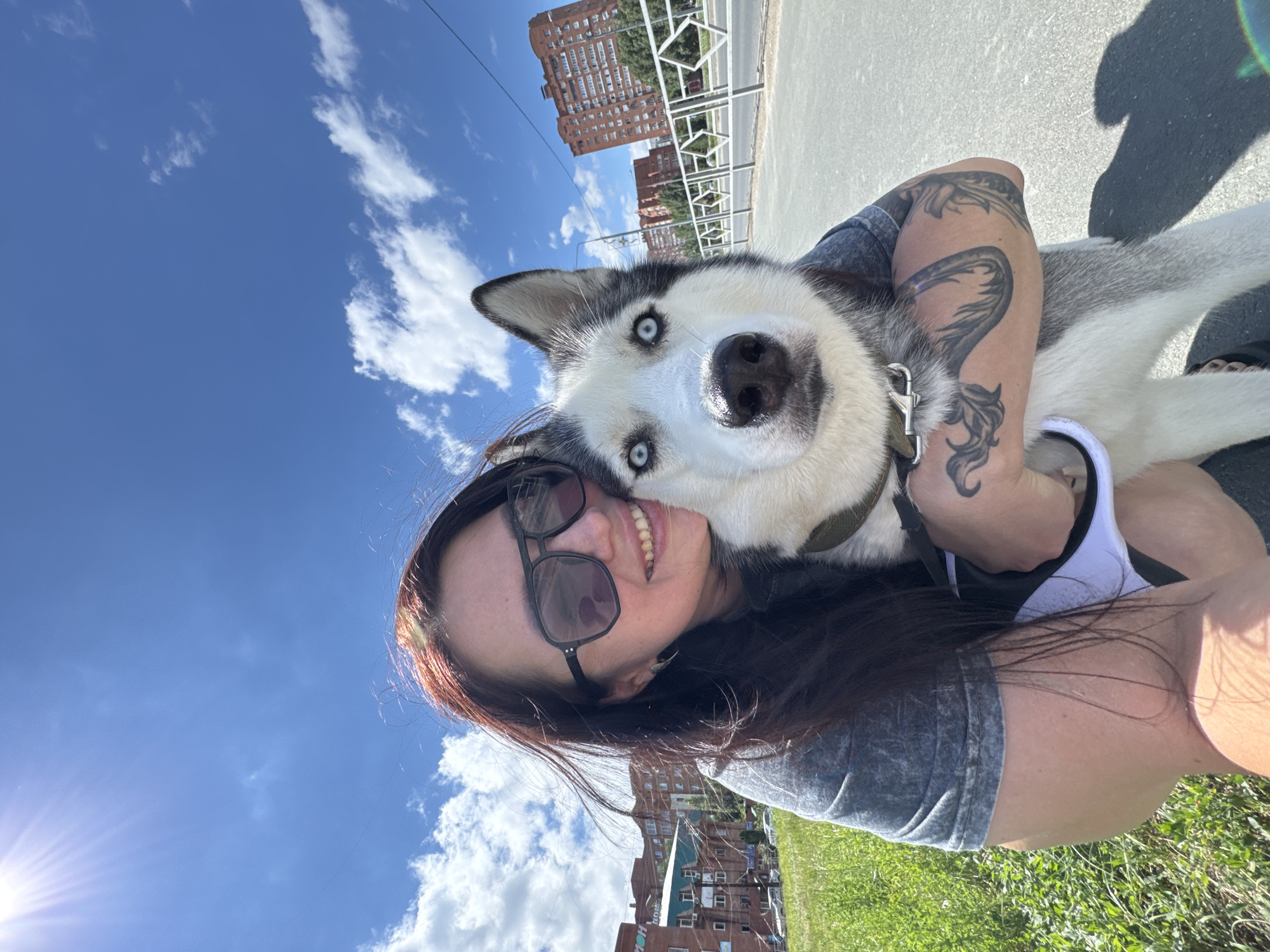 A woman with dark hair and sunglasses takes a selfie outdoors, holding a husky dog. The dog has striking blue eyes and a black and white coat. They are standing on a paved path, with a grassy area to the left and a street with cars and buildings in the background. The sky is a bright blue with scattered white clouds and the sun is shining, indicating a warm day. The buildings in the background are typical of urban residential areas, some appearing to be apartment blocks. The overall scene suggests a casual outing in a city park or on a sidewalk. The presence of buildings and a paved road indicates an urban environment. The specific location in Tolyatti, Russia, is not directly ascertainable from the image itself but can be inferred from the context.