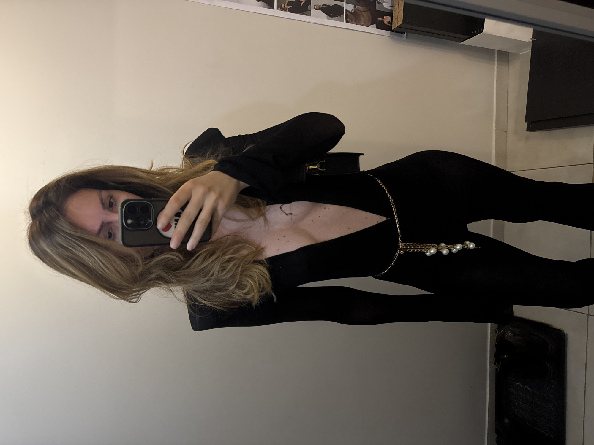 A woman is taking a selfie in what appears to be a changing room or a small room with a mirror. She is wearing a black, long-sleeved jumpsuit with a plunging neckline, a gold chain belt with pearl-like embellishments, and a small black purse over her shoulder. Her hair is long and wavy. The background is a plain, light-colored wall. To the left, there is a dark mat on the floor with what appear to be black shoes. To the right, there are some framed images on the wall, suggesting it might be a retail space or a dressing room within a store. There is no indication of the city of Sofia, Bulgaria, in the visual elements of the image.