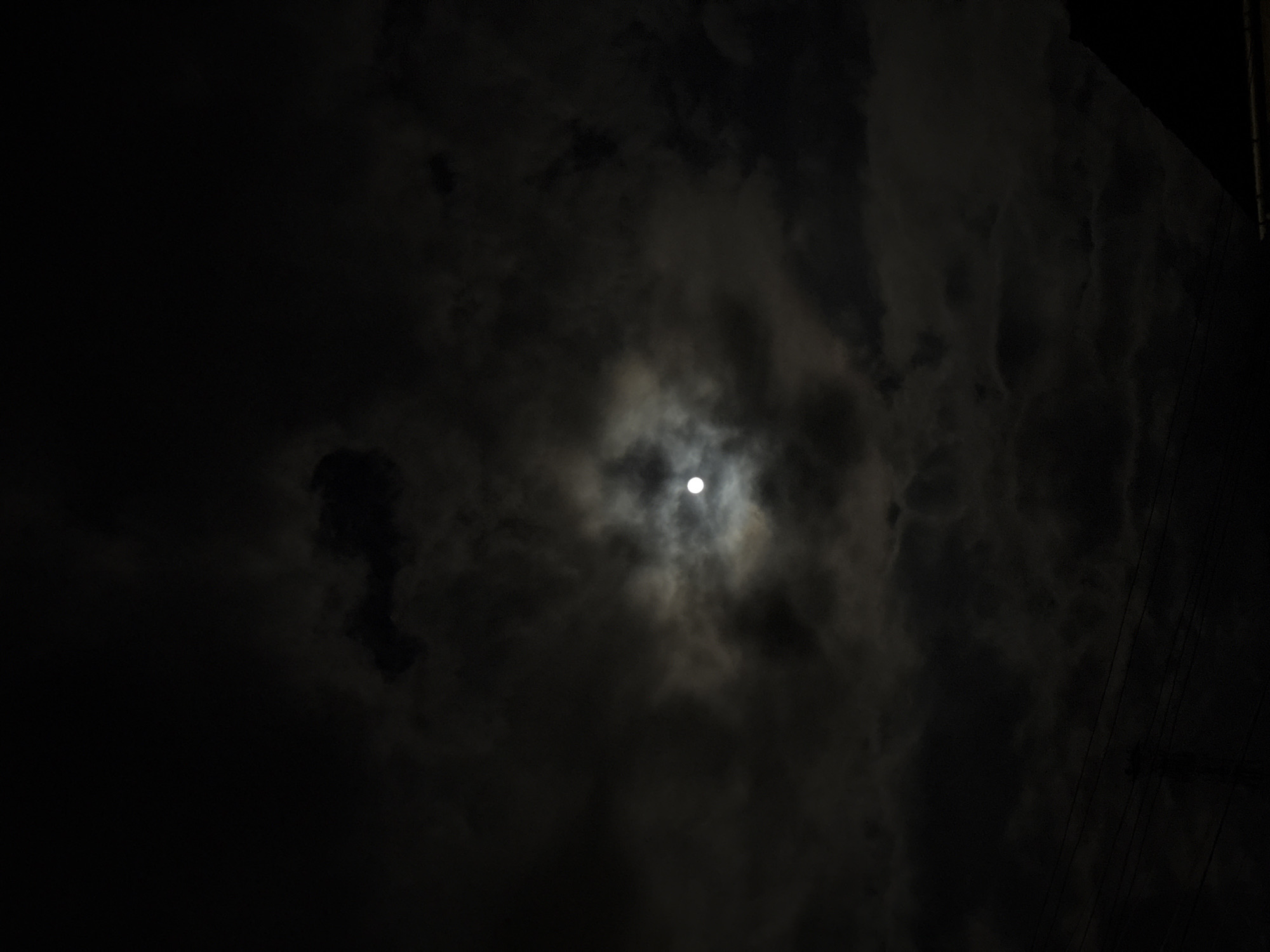 The image depicts a night sky with a full moon obscured by clouds. The moon is visible as a bright, round spot surrounded by a halo of lighter clouds, contrasting with the darker areas of the sky. Silhouettes of power lines and a corner of a building can be seen in the upper right corner of the image. No people are visible. The photograph was taken in Kozloduy, Bulgaria.