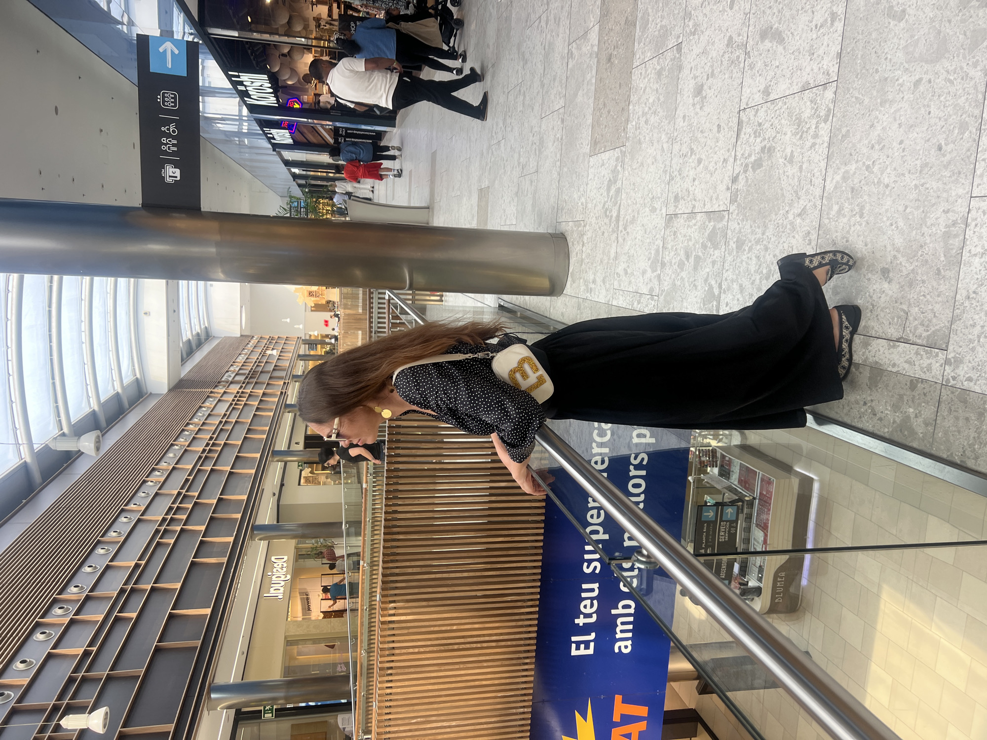A woman stands near a railing in a shopping mall. She wears a polka-dot blouse, a black skirt, and flats. She has a purse with a large "G" on it. The mall has multiple levels, with stores like "Desigual" and "Koishi" visible. Signage includes directions and ATM indicators, with text in Catalan ("El teu supermercat"). The floor is tiled in a light grey pattern. The location is the Parc Central shopping mall in Tarragona, Spain.