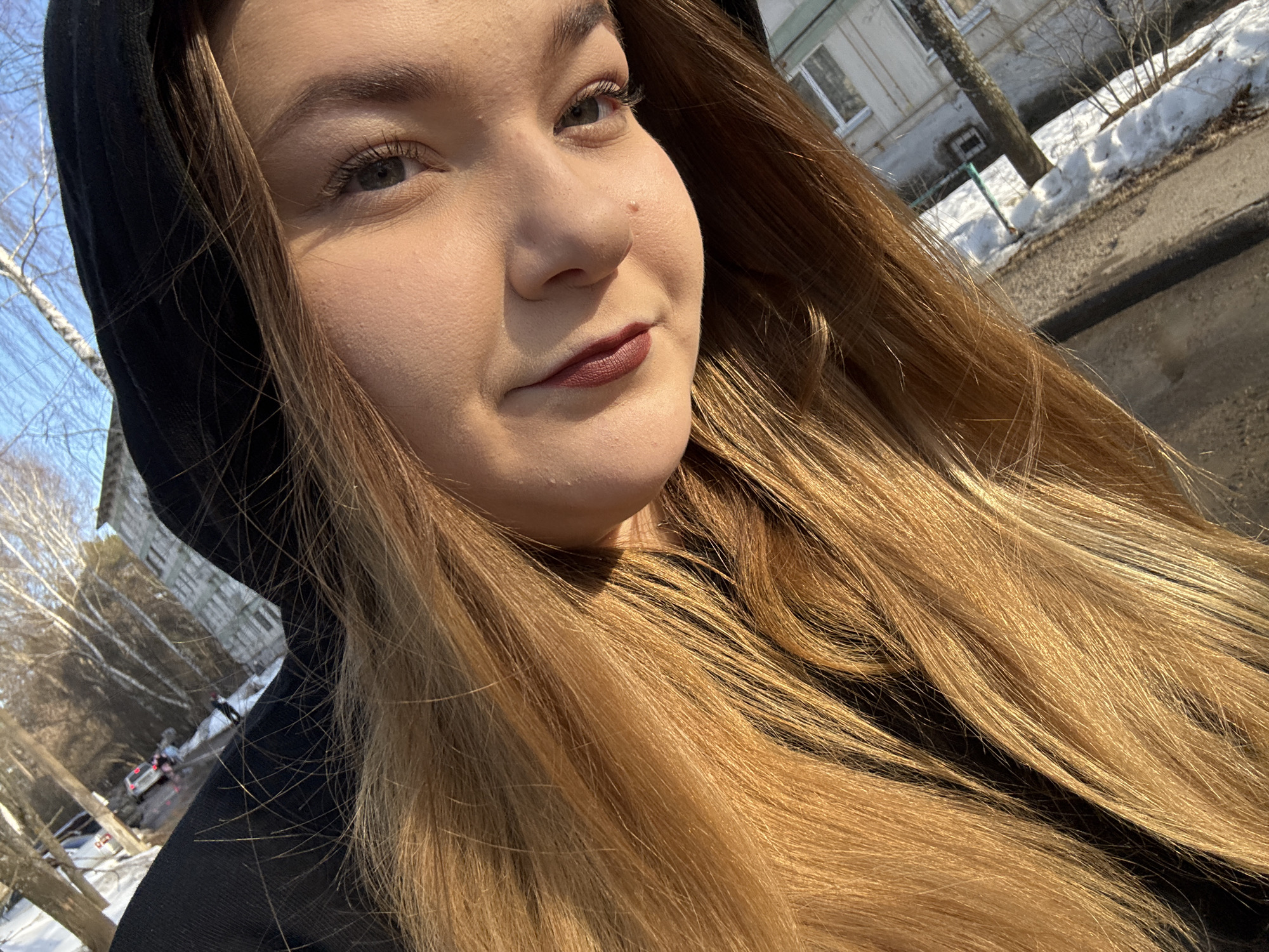 A fair-skinned woman with light brown hair and wearing a black hooded sweatshirt is taking a selfie outdoors. The background suggests an urban environment with remnants of snow, trees, buildings, and a road. Based on the metadata, this photo was taken in Chaikovsky, Russia.