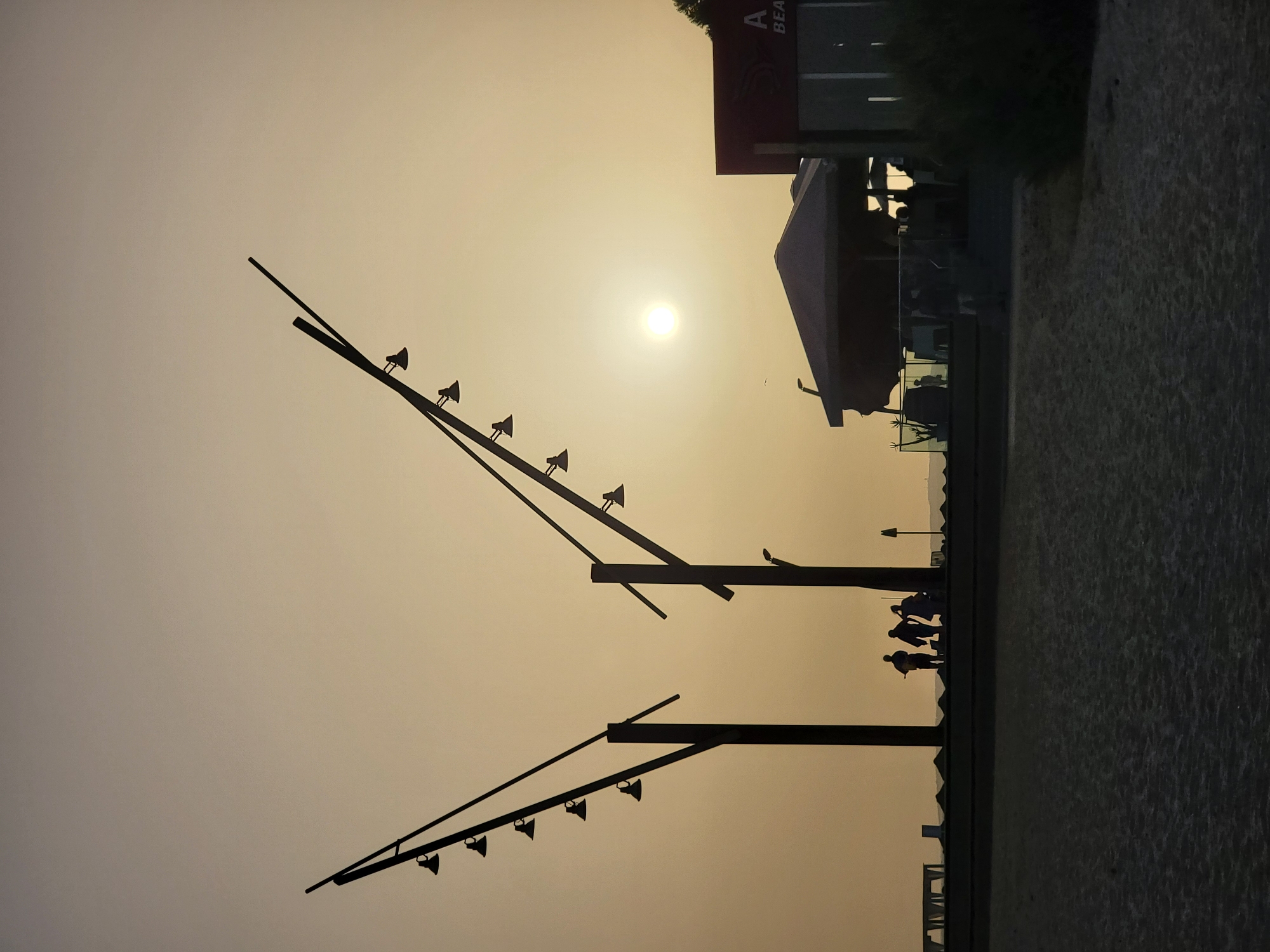 The image depicts a silhouetted scene at dusk on a beach in Quarteira, Portugal. In the foreground, the dark sand of the beach stretches towards a raised walkway. Along the walkway, several figures are silhouetted against the hazy, sunlit sky. Two large, V-shaped lampposts with multiple lights are prominent on the left side of the frame. To the right, a building with a red sign and an awning is visible, also in silhouette. The sun is a bright, hazy orb in the sky, suggesting a low horizon. The overall atmosphere is tranquil and the lighting is soft, characteristic of a beach at sunset.