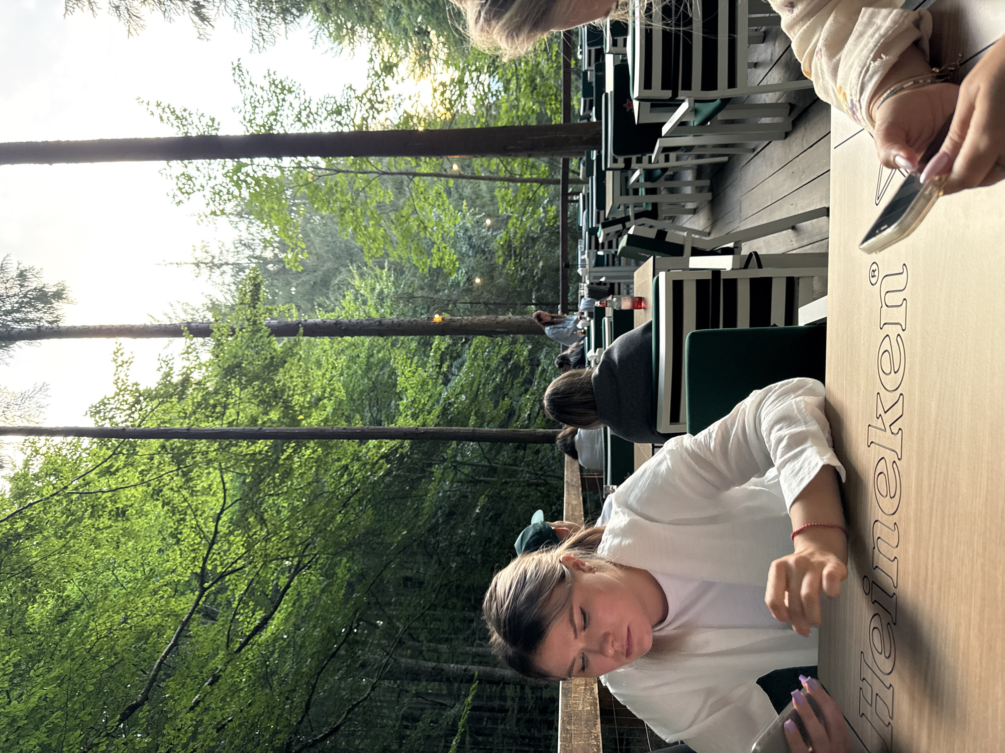 The image depicts an outdoor seating area of a restaurant nestled within a dense forest, likely in Arefu, Romania, given the surrounding foliage and towering trees which are characteristic of the area. The foreground features a young woman, appearing to be in her late teens or early twenties, seated at a wooden table. She is looking down at her smartphone, which she holds in her left hand. Her right hand is resting on the table. She is wearing a white, loose-fitting shirt. The table surface has the embossed logo "Heineken".
In the background, several other individuals are visible seated at tables, suggesting a leisurely dining or social gathering. The seating area is comprised of numerous tables and chairs arranged on a wooden deck. The chairs are primarily white with dark green cushions. The environment is rich with lush green trees and foliage, with sunlight filtering through the canopy, indicating daytime. The overall atmosphere appears relaxed and natural, with the restaurant integrated harmoniously into the forest setting. The focus of the image is on the woman in the foreground and her interaction with her device, juxtaposed against the serene natural backdrop.