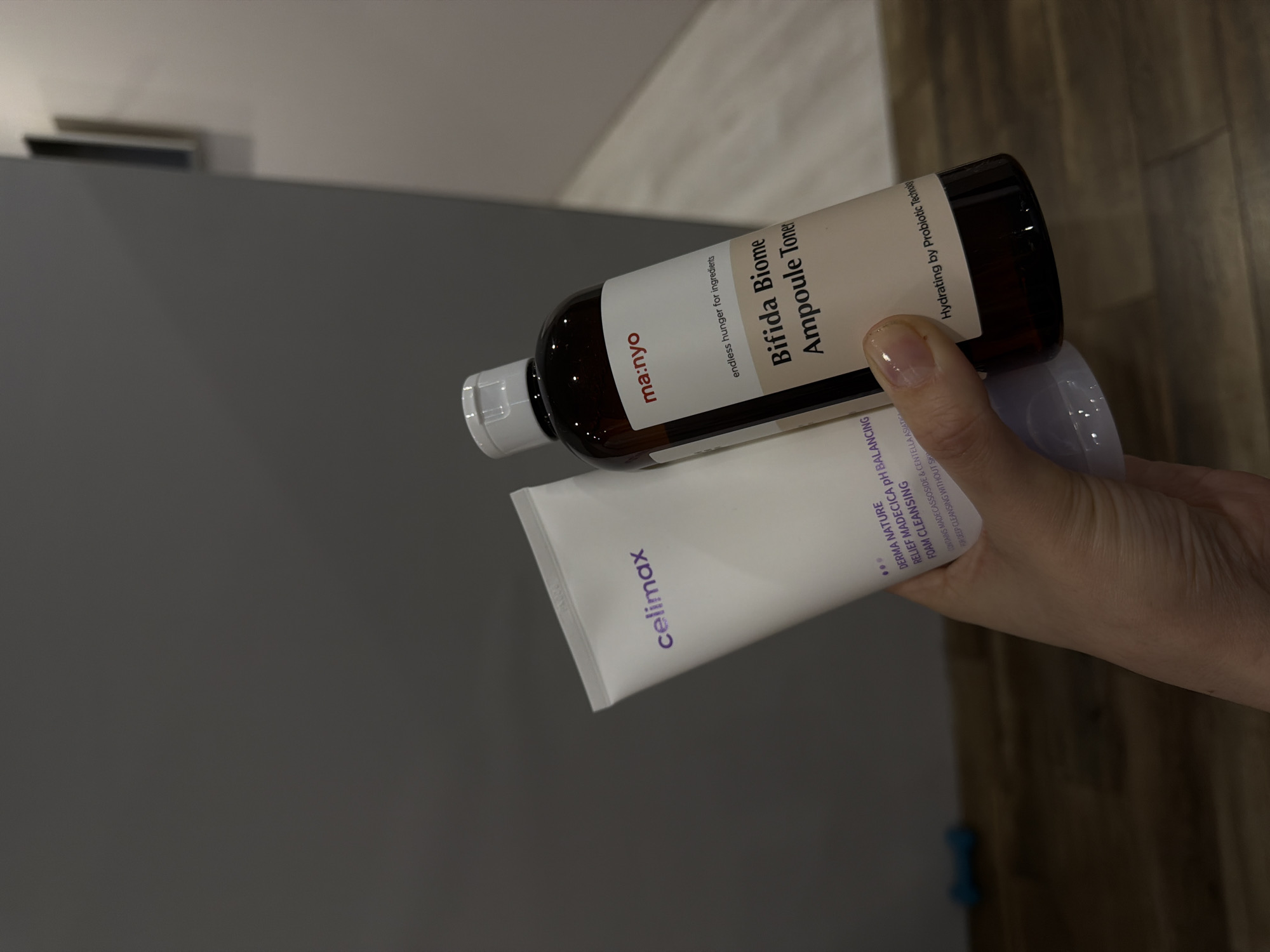 A hand holds a Celimax Derma Nature Relief Madecica pH Balancing Foam Cleansing tube and a Ma:nyo Bifida Biome Ampoule Toner bottle against a neutral background. The bottle is brown with a white label featuring product information, while the tube is white with purple Celimax branding and product details. The hand is slightly visible, and the background includes a gray wall and a wooden surface. The location of Chaikovsky, Russia, is not evident from the image.