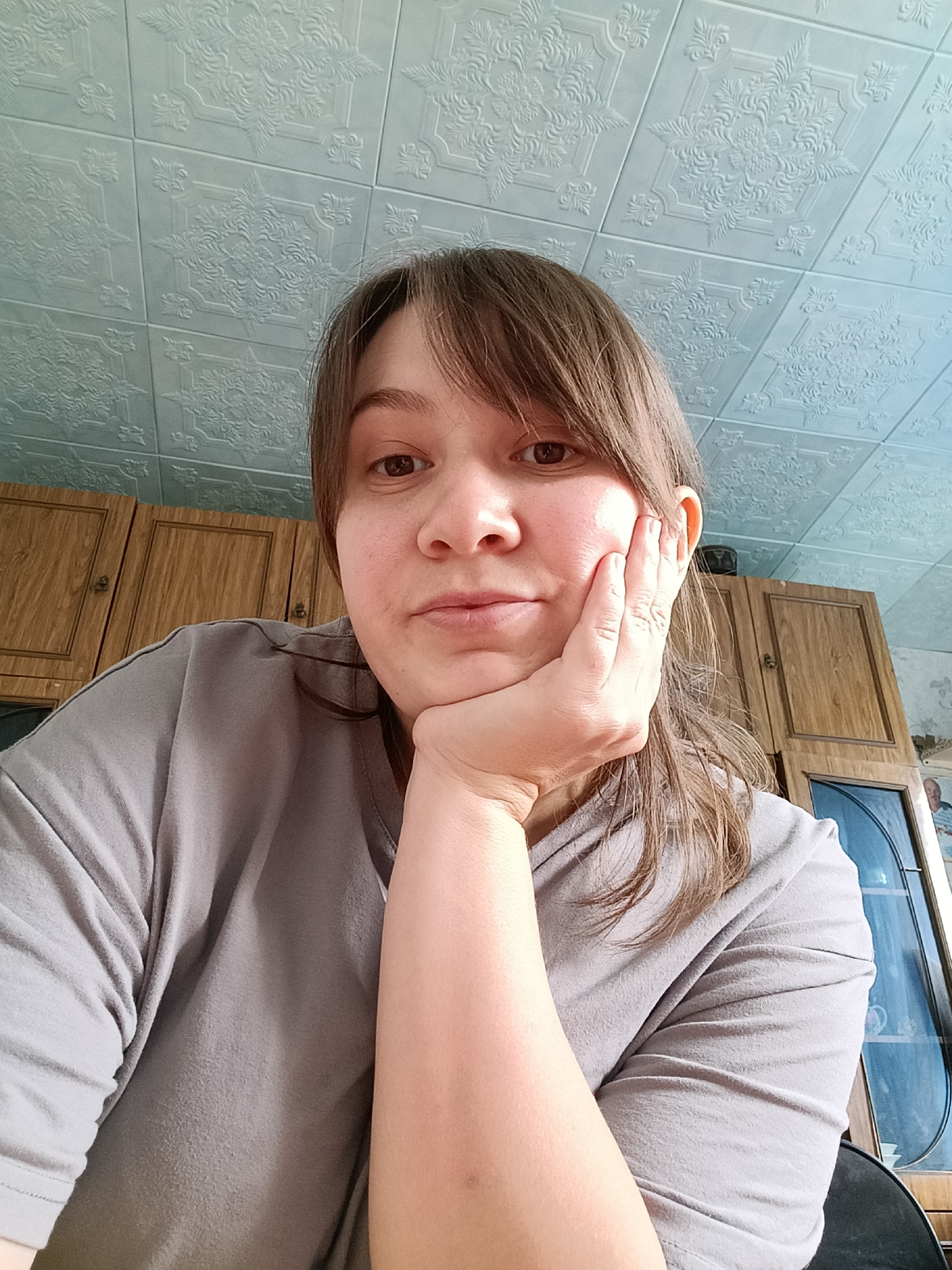 A medium shot photograph taken from a high angle captures a young woman with light brown hair and bangs resting her chin on her left hand, her gaze directed towards the camera. She is wearing a plain grey t-shirt. The background consists of a light blue ceiling with an embossed patterned texture and wooden cabinets with dark knobs. To the right of the woman, a glass-fronted cabinet is visible, containing items on its shelves. The overall setting appears to be an indoor domestic environment, likely a kitchen or dining area, within the city of Каменное, Russia.