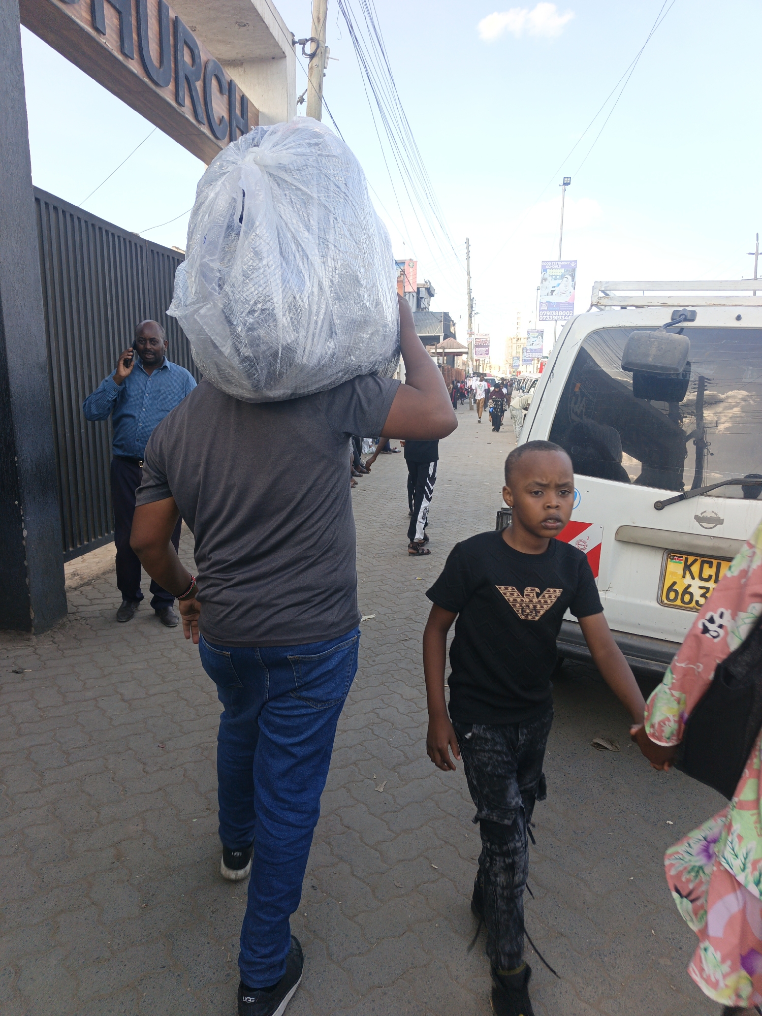 The image captures a street scene in Nairobi, Kenya. A man is carrying a large, clear plastic bag over his shoulder, walking away from a building labeled "Church." He is wearing a grey t-shirt, blue jeans, and black sneakers with "UGG" written on the back. To his right, a young boy in a black t-shirt with a logo and grey patterned pants is walking with someone whose floral pink dress is partially visible on the right. A white van with a Kenyan license plate "KCI 663" is parked on the right side. A man in a blue button-down shirt is talking on a cell phone near the church entrance. Other pedestrians and vehicles are visible in the background, along with street signs and banners. The street is paved with grey bricks. Power lines are above the scene.