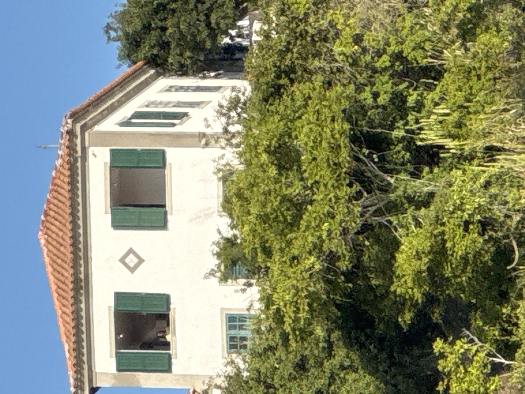 The image shows a two-story white building with an orange tiled roof, situated on a hill covered in lush green foliage. The building has several windows, some with green shutters. One window on the upper floor shows a person with light-colored hair wearing a yellow top. Another window on the upper floor is open. Below the upper windows, there is a diamond-shaped decorative element. On the lower floor, there is a window with blue-tinted panes and green shutters to its right. A large amount of dense green vegetation covers the foreground and the base of the building. The sky is a clear blue. The building's architectural style suggests it could be in a historical or Mediterranean-influenced region. Without additional context, it is not possible to confirm its specific location in Niterói, Brazil, as the image itself does not provide any identifying landmarks or signs.