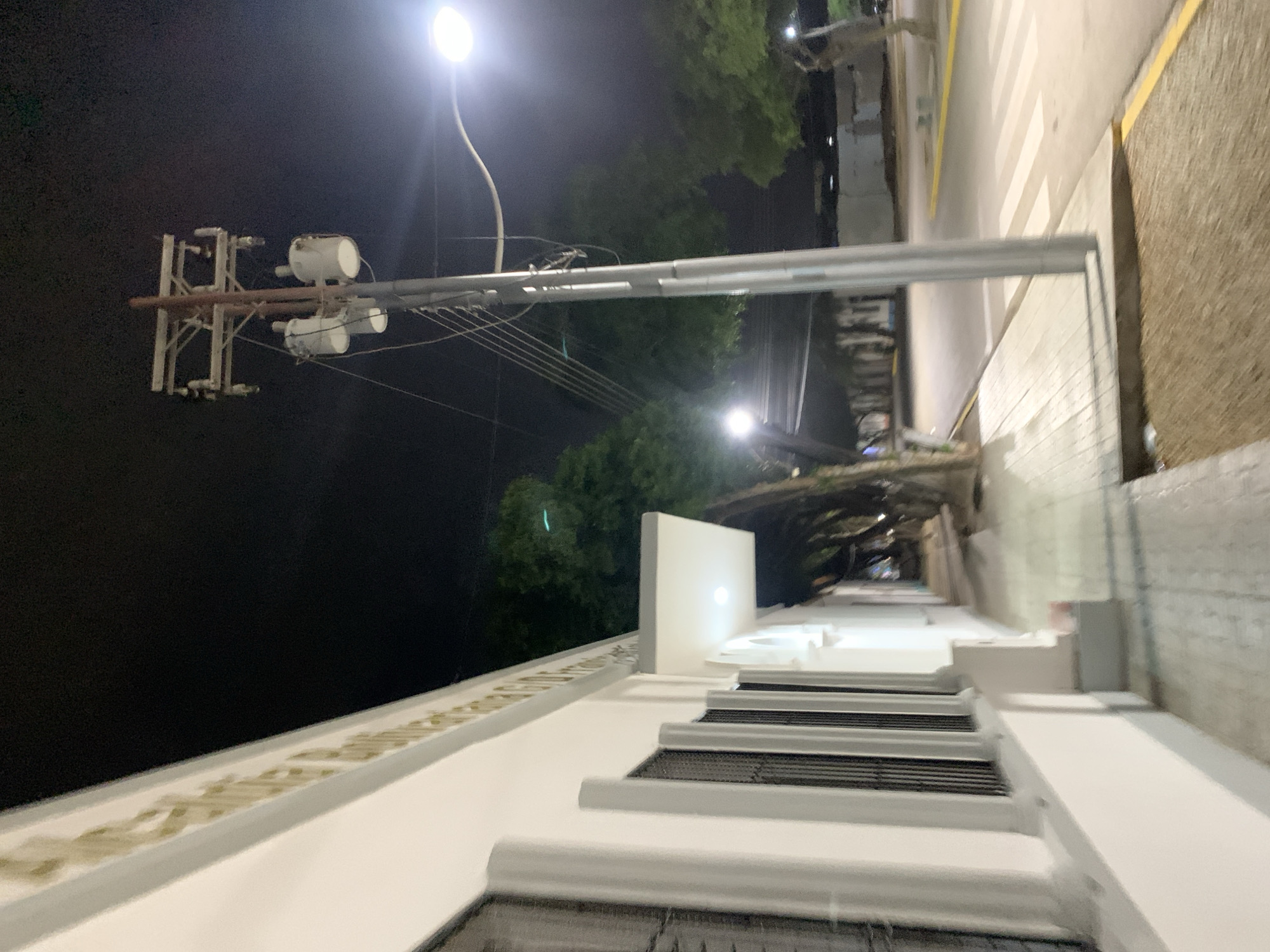 The media file presents an elevated, nighttime view of a street in Santa Ana, Venezuela. The scene is illuminated primarily by artificial lights, with a dark sky overhead.
In the immediate foreground, the white facade of a building is visible, featuring distinct architectural details that resemble stacked steps or ledges with dark, possibly grated, openings. Along the top edge of this white structure, golden-colored text is partially discernible. Examining the cropped image, the text clearly reads "SANTA AN...", strongly suggesting a reference to "Santa Ana."
Across the street, a prominent utility pole rises into the dark sky. It is equipped with multiple crossarms supporting a complex array of wires and several white, cylindrical pieces of equipment, which could be cellular antennas or other communication devices. A long, light-colored horizontal beam or conduit also stretches across the mid-ground, parallel to the utility pole. Two bright streetlights are visible; one is particularly intense and centrally located, while another can be seen further down the street. Dark green foliage from trees is interspersed throughout the background against the night sky.
The street below is paved and features white crosswalk markings, along with yellow lines delineating the curb. A sidewalk runs alongside the road. The overall impression is one of a quiet, urban or semi-urban environment at night, devoid of people or visible activity. The clear night sky and the functional infrastructure are well-lit by the streetlights.