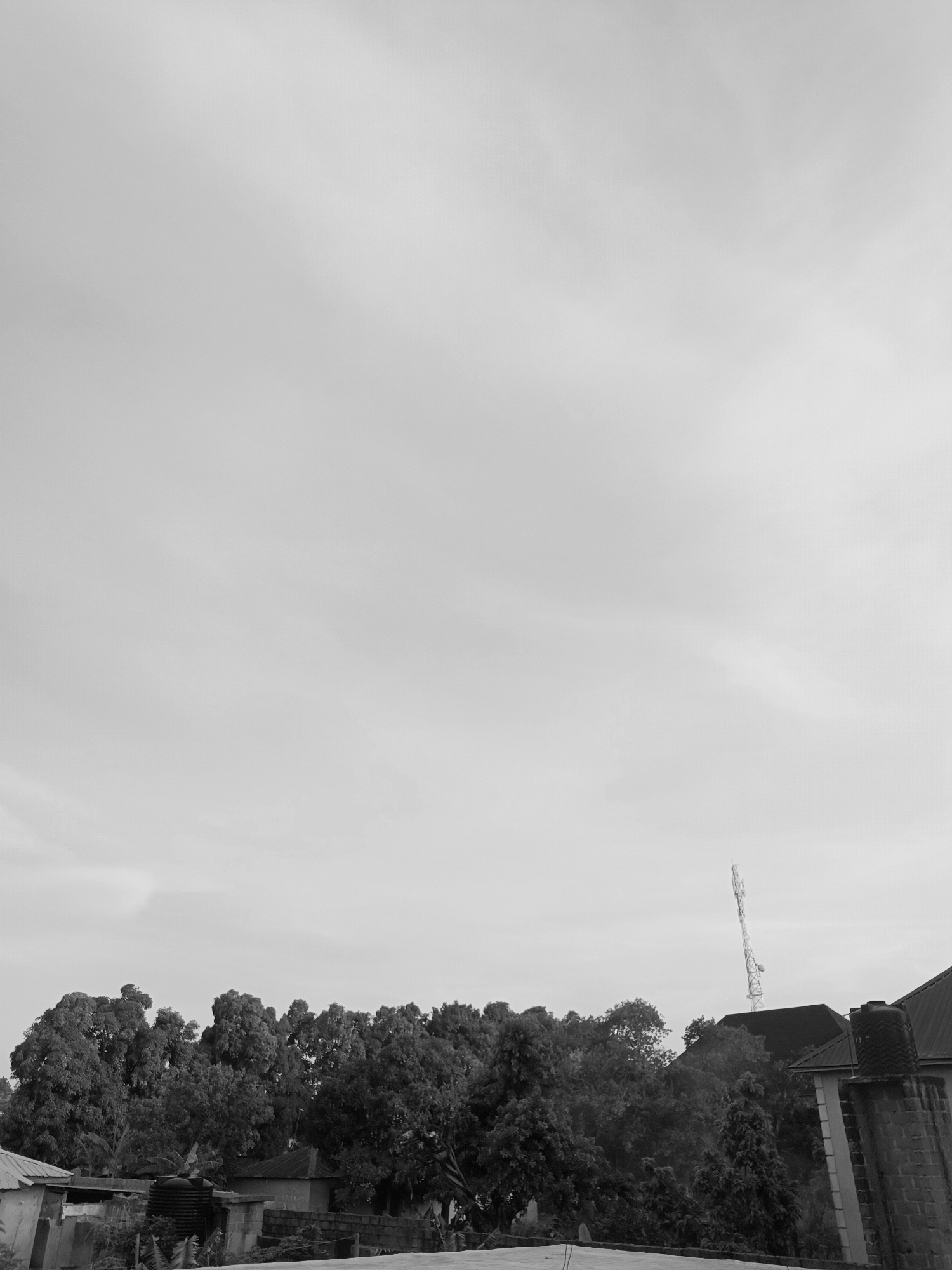 The image, taken in Bukuru, Nigeria, is a grayscale medium shot with the sky taking up the majority of the frame. Below the sky is a line of trees and rooftops of buildings. A tall cell phone tower is on the right side of the image, behind the trees and buildings. There are no people in the image.