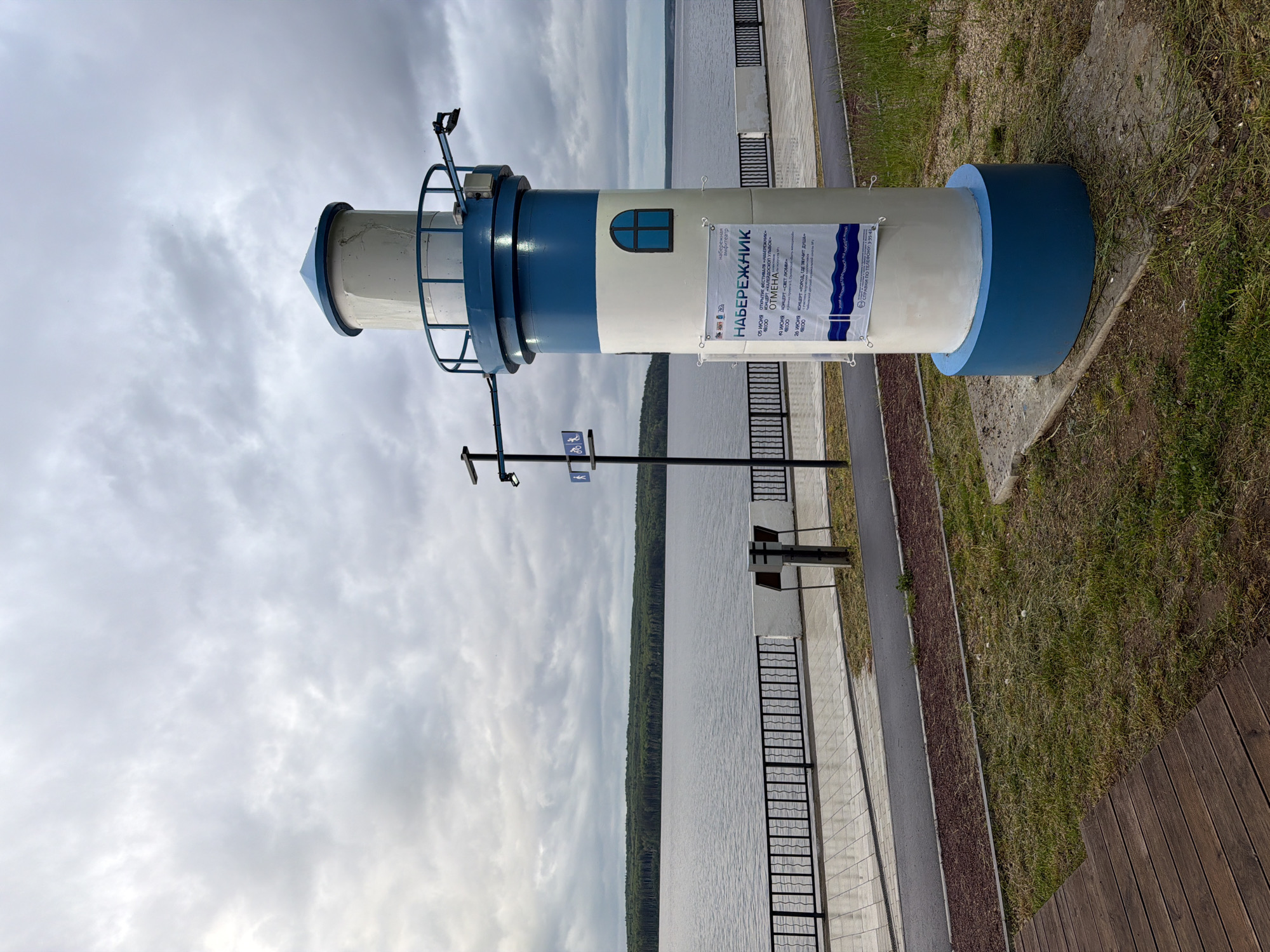 This image depicts a waterfront scene in Chaikovsky, Russia, characterized by a large body of water under a heavily overcast sky. The foreground features a decorative, miniature lighthouse positioned slightly to the right of the frame, painted in alternating bands of white and blue. Its base is blue, followed by a white cylindrical section with a small, arched, dark-framed window. A blue band encircles its middle, topped by another white section with a blue-railed observation deck and a blue conical roof. An arm extends horizontally from the top section to the right, possibly holding an outdoor light or sensor. Several white posters with blue text, written in Russian, are affixed to the lighthouse body. The top poster displays the title "НАБЕРЕЖНИК" (Embankment) and lists event dates, with one event explicitly marked "ОТМЕНА" (CANCELED).
The lighthouse stands on a patch of grass, with a dark brown wooden boardwalk partially visible in the bottom-left corner. Immediately behind the grass and the lighthouse is a paved promenade. This promenade features light-colored tiles and a darker asphalt path, separated from the water by a black metal fence with wavy top elements, backed by a low white wall. To the left of the lighthouse, on the promenade, a dark grey informational stand or kiosk is visible. Further left, a utility pole displays two white-on-blue signs, one depicting a walking person and another a bicycle, indicating a pedestrian and cycling path. The background is dominated by a wide, calm body of water, likely the Kama River or Votkinsk Reservoir, extending to a distant horizon marked by a dense line of dark green forest. The sky is uniformly grey with thick clouds, suggesting an overcast day with muted light. No people are visible in the scene.