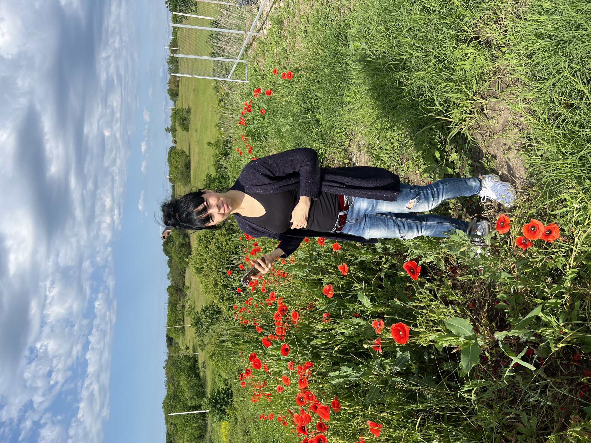 A woman stands in a field of red poppies in Tugurgoy, Russia. She wears a black top and cardigan, ripped blue jeans, and white sneakers. She is holding a cellphone and appears to be taking a photo. The sky is blue with scattered clouds. A green field with a fence is visible in the background.