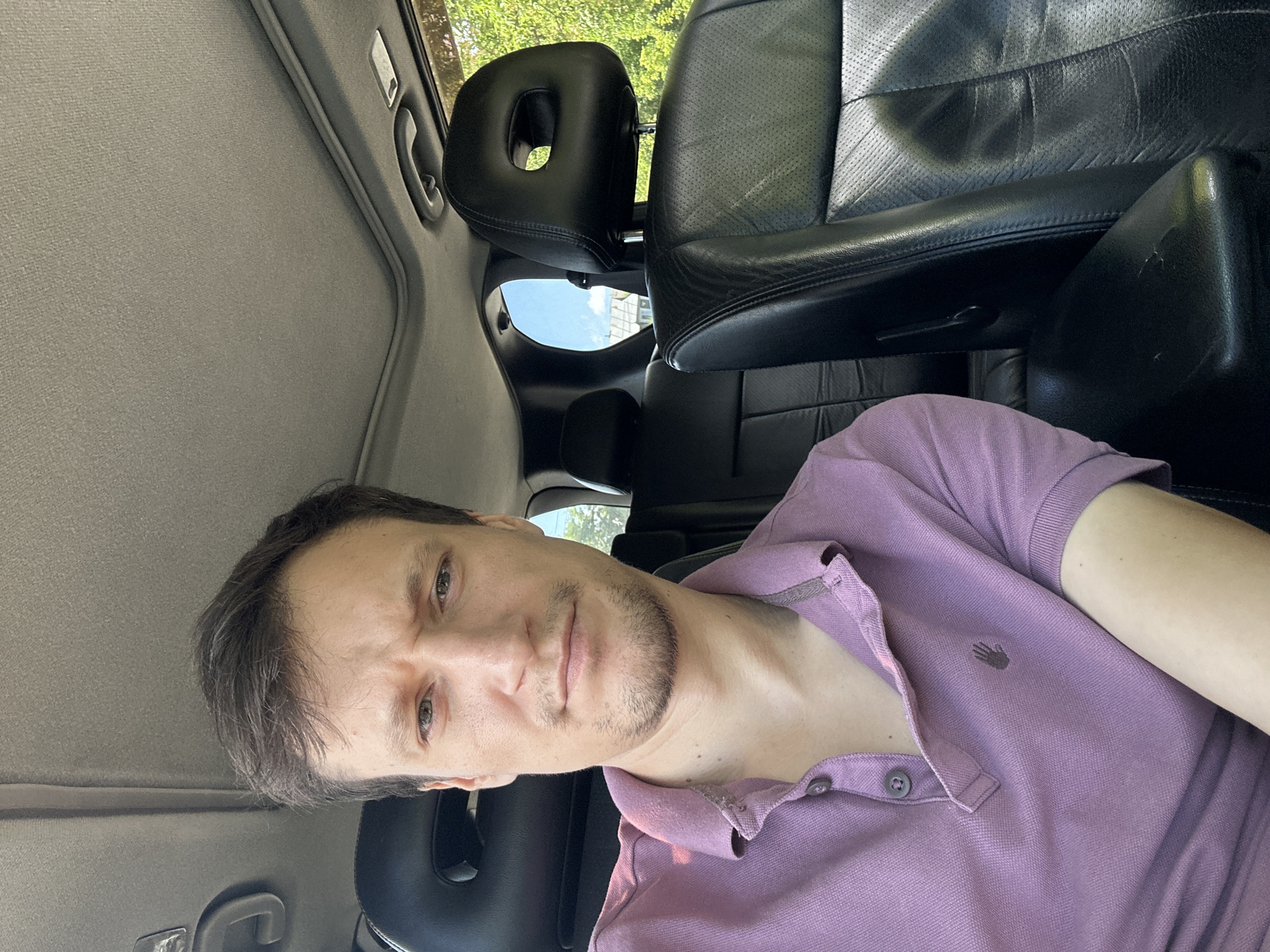 A medium shot captures a man from the chest up, seated in the passenger seat of a car. He has light brown hair, fair skin, and a short beard. He is wearing a purple polo shirt with two buttons at the neck and a small, embroidered hand logo on the left side of his chest. The car's interior is visible, with black leather seats, a sunroof, and a window showing green trees and a blue sky. The man is looking directly at the camera with a neutral expression. The location is not specified in the image or context.