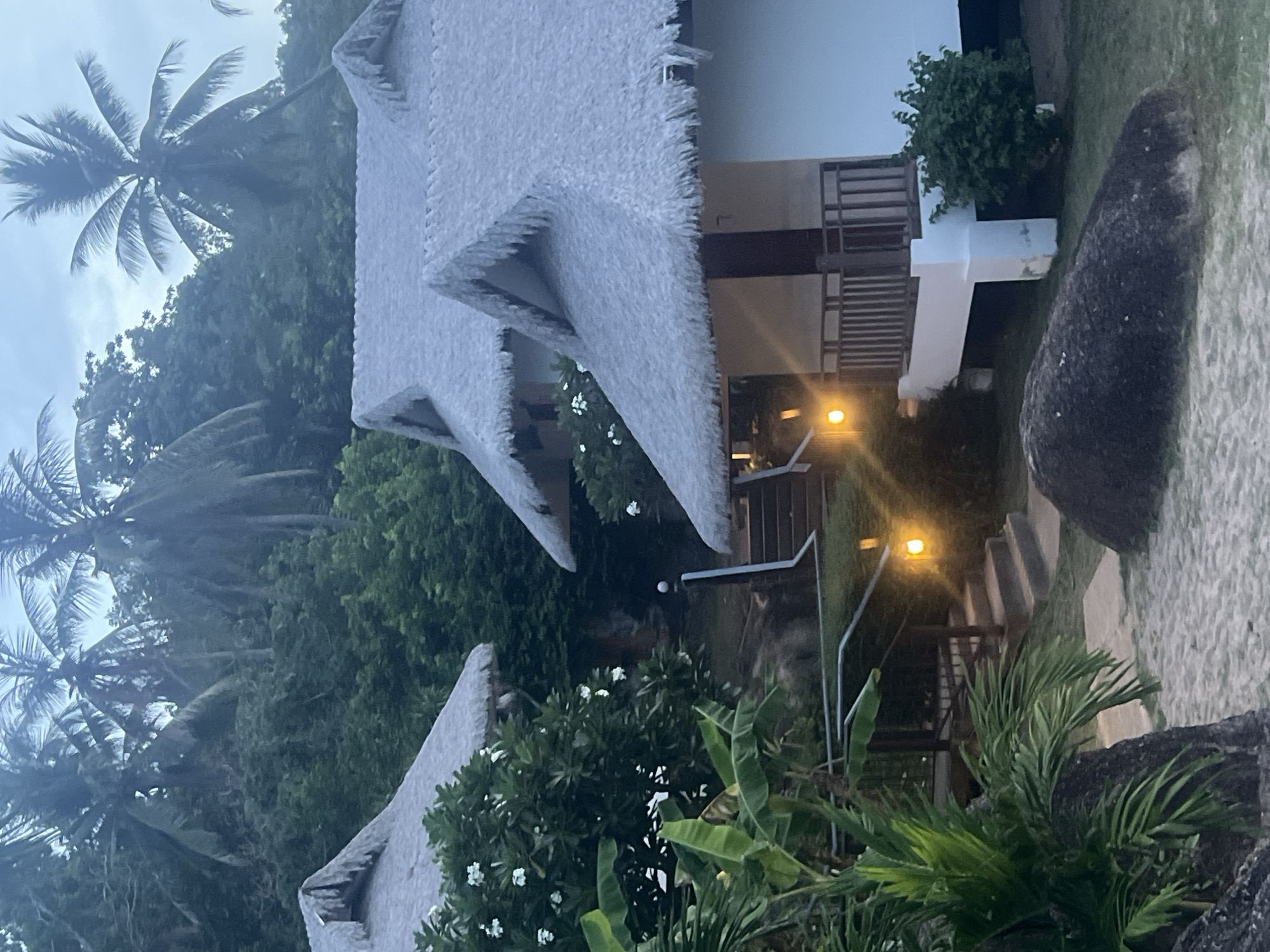 The image depicts a serene scene in Ko Samui District, Thailand, likely at dusk. Several thatched-roof bungalows are nestled amongst lush green foliage, with towering palm trees dominating the background. The bungalows feature white walls and dark wooden accents, with small balconies and stairs leading up to them. Soft yellow lights illuminate the pathways and stairs, creating a warm and inviting atmosphere. Large, dark boulders are scattered on the sandy ground in the foreground, adding to the natural, tropical ambiance. The sky is overcast, suggesting a calm and peaceful evening. No people are visible in the image, focusing the viewer's attention on the tranquil architecture and the surrounding natural beauty.