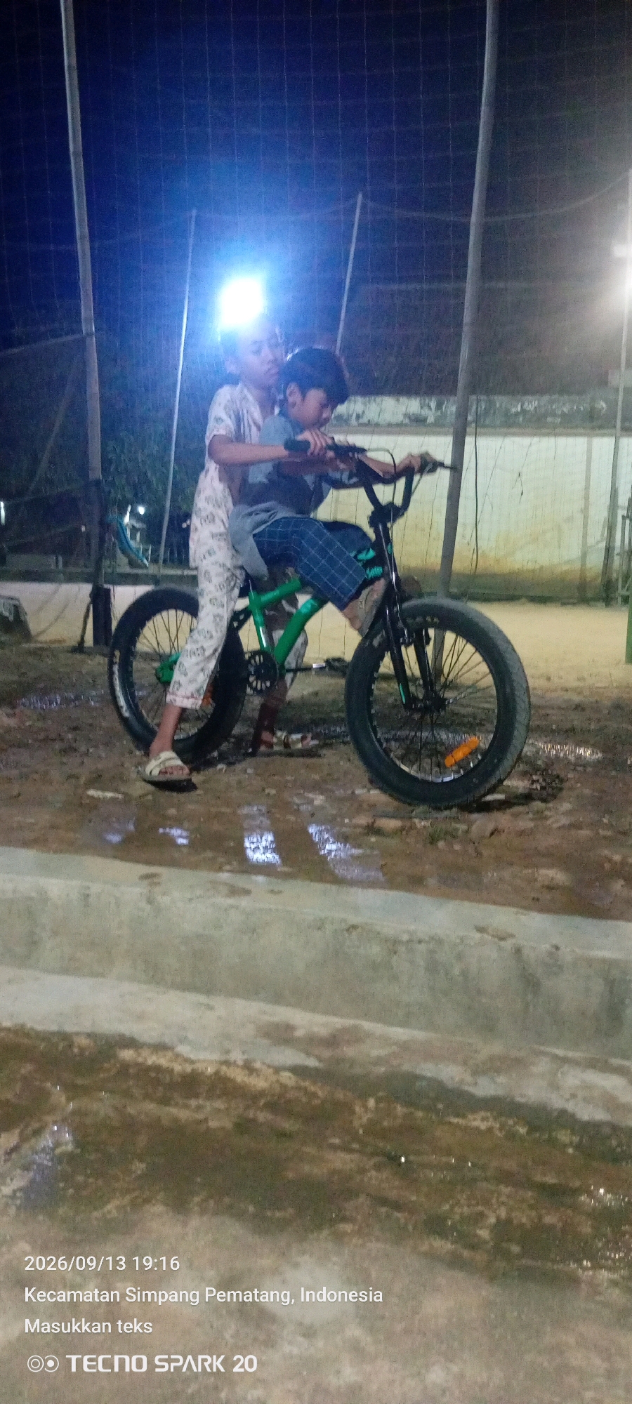 In Simpang Pematang, Indonesia, two children are captured in an evening outdoor setting, illuminated by artificial lights. A younger child, wearing a blue t-shirt and plaid shorts, is seated on a green and black fat-tire bicycle, operating the handlebars. An older child, dressed in light-colored pajamas, stands behind the seated child, their arms positioned around the older child's torso. The ground appears wet and muddy, indicating recent rain or a damp environment. The background shows a net-like structure, possibly a sports field enclosure, and a light-colored wall. The photograph was taken on September 13, 2026, at 19:16, and the image contains the watermark "TECNO SPARK 20".