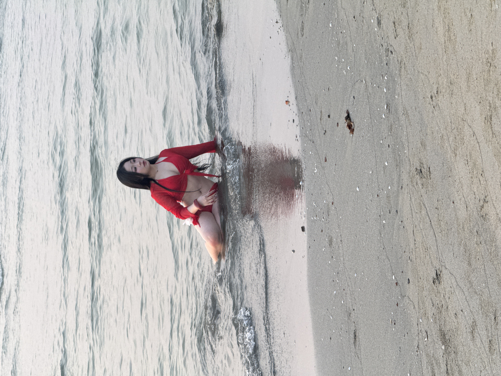 A woman wearing a red long-sleeve top and shorts sits in the shallow water at the edge of a sandy beach. The location is likely a beach in the coastal city of Đà Nẵng, Vietnam, given the landscape and geographic location. The woman's legs are immersed in the water, while her upper body is partially on the sand. The water is slightly choppy, with small waves breaking on the shore. The sand is light-colored and appears to be fine-grained. The image is taken from an angle that shows both the woman and the surrounding beach environment.