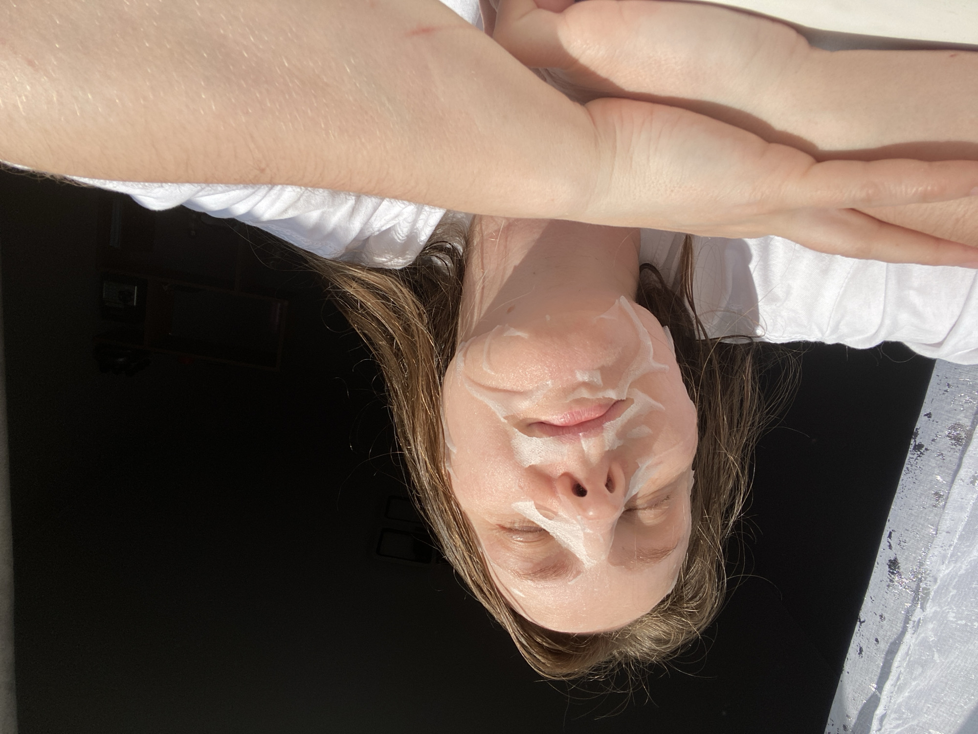 A person is pictured with a white facial mask on their face. Their eyes are closed. Their skin is fair, and they have brown hair. They are wearing a white top. An arm and hand are visible in the upper portion of the image. The background appears to be dark. The image is rotated, placing the subject upside down. Location information, specifically the city of Noviy, Russia, is not identifiable within the frame.
