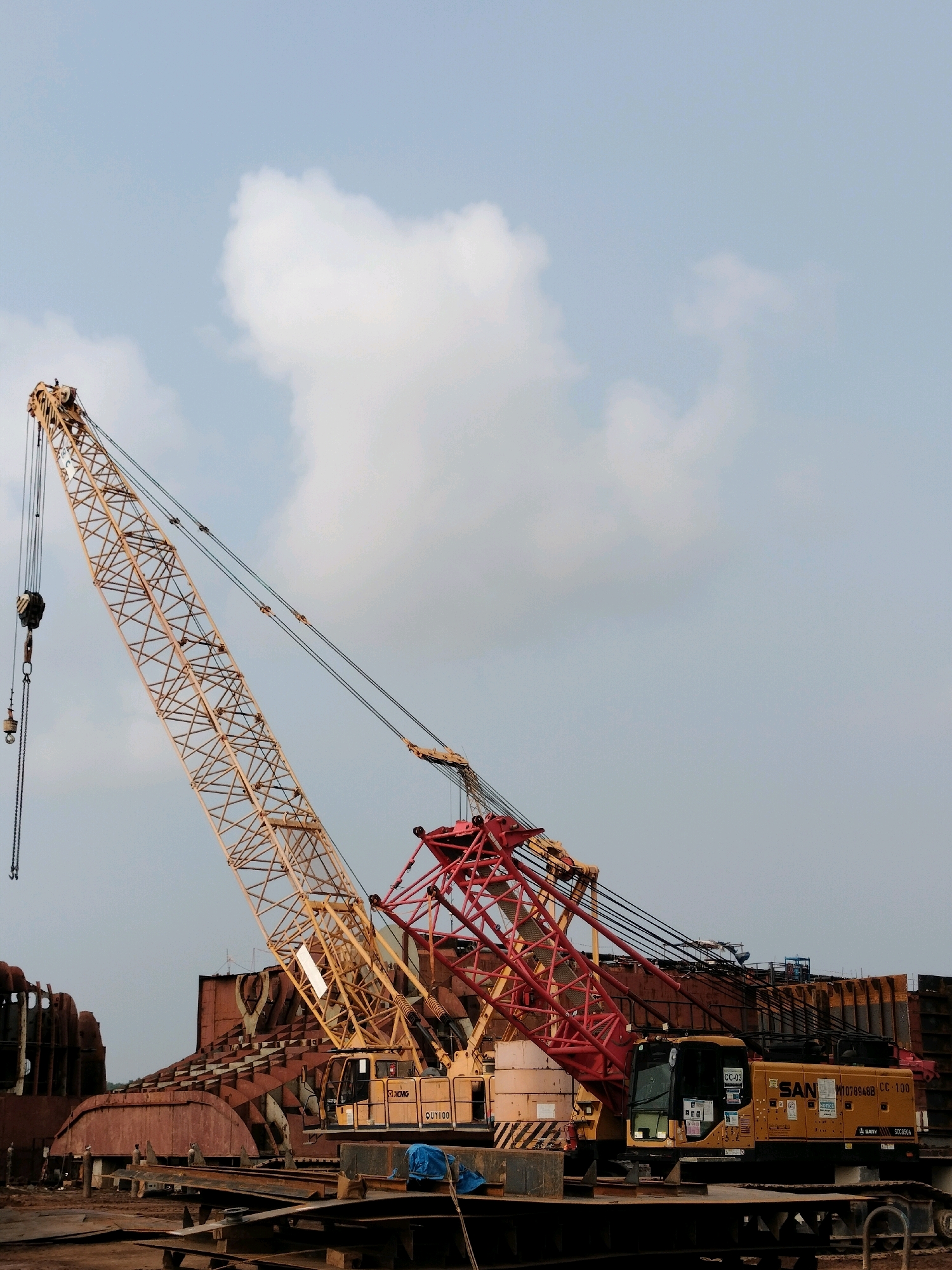 The image depicts a shipyard scene in Batam, Indonesia, focusing on large industrial cranes and the construction of a massive vessel. In the foreground, a yellow mobile crane, identified by the markings "SANY CC-100" and model number "1076948BB", is positioned on a platform. To its left, a taller, yellow lattice boom crane dominates the frame, with its jib extending high into the sky. Behind these cranes, the skeletal structure of a large ship under construction is visible, characterized by its rusty, curved metal plates and internal supports. The environment suggests an active construction site, with various metal components and equipment scattered around the ground. The sky above is partially cloudy, with a light blue hue. The scene is static, capturing a moment of industrial activity.