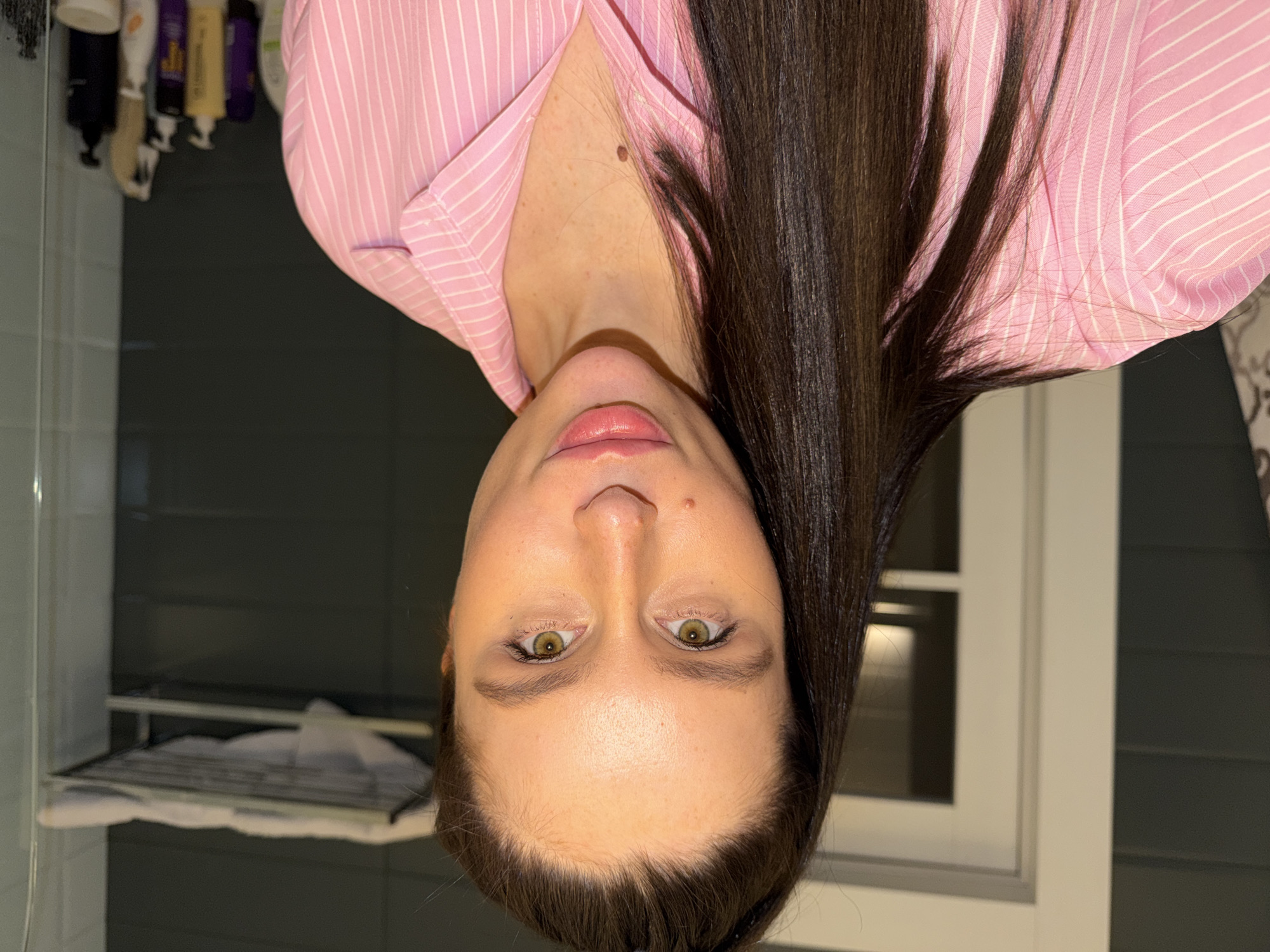 This close-up portrait features a young adult woman looking directly at the camera. Her head is positioned centrally, slightly tilted, with long, dark brown hair cascading over her shoulders and chest. She is wearing a pink and white striped button-up shirt, partially unbuttoned. Her eyes are light-colored, possibly green or hazel, and her lips are full with a subtle pink gloss. A small mole is visible on her upper chest/neck area.
The background suggests an indoor setting, likely a bathroom, in what is stated as Khimki, Russia. To the viewer's left, partially obscured by her hair, is a section of a dark grey or black wall and a clear shower glass panel. Above her left shoulder, several pump bottles and other toiletries are neatly arranged on a shelf. To the viewer's right, a window with white framing is visible against a similar dark wall, and below it, a metal towel rack holding white folded towels can be seen. The lighting appears to be artificial and even. The woman's expression is neutral and calm. There is no clearly legible text in the image.
