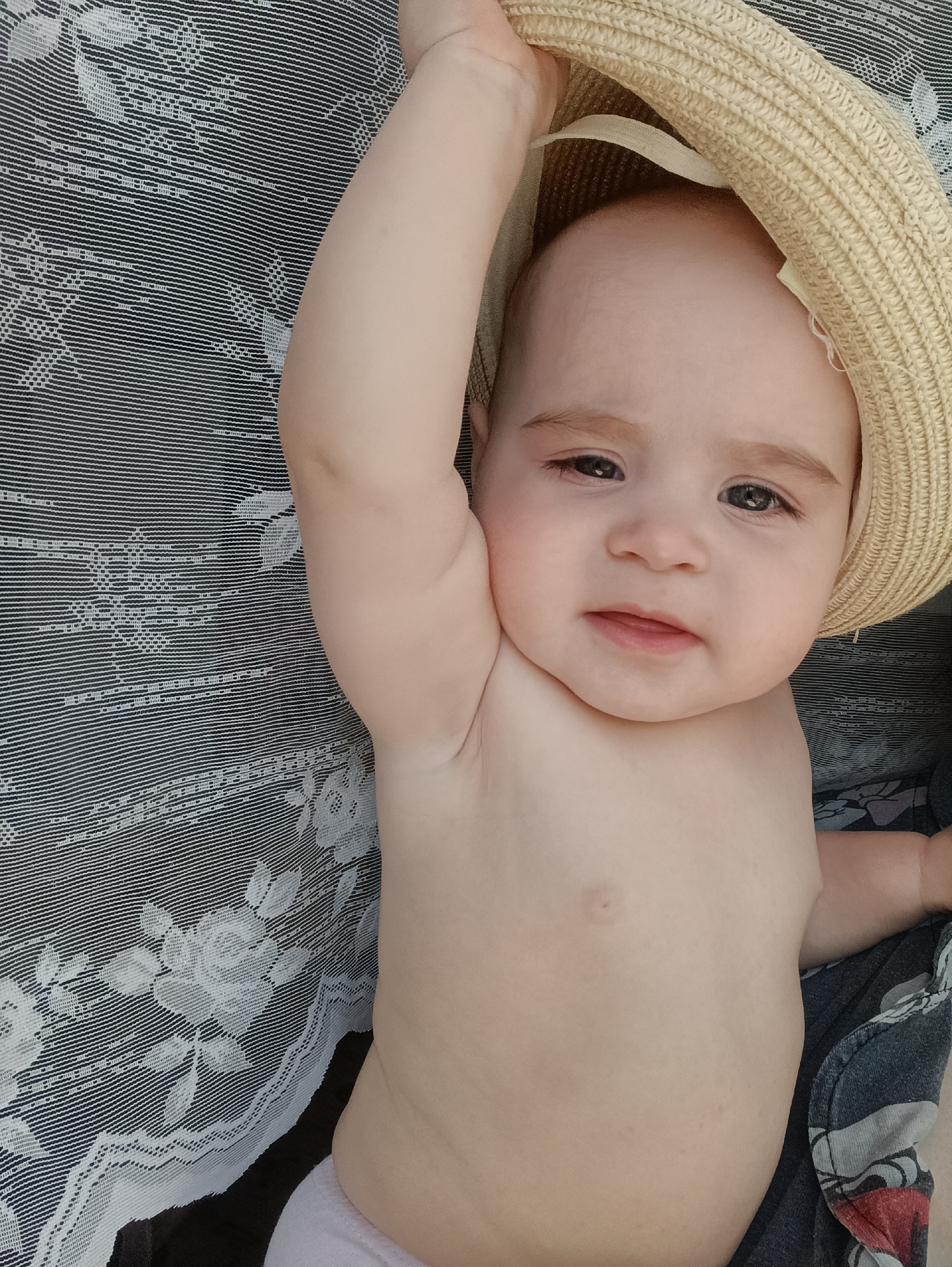 A baby lies on a black and white floral patterned blanket wearing a straw hat. The baby's right arm is raised, and the hat is partially obscuring the baby's face. The setting is unknown.