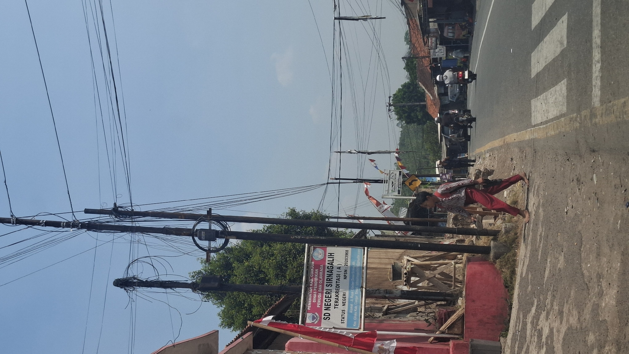 A street scene in Sirnagalih, Indonesia, unfolds under a clear blue sky, dominated by numerous electrical wires crisscrossing overhead, supported by wooden utility poles. In the foreground, a young boy walks barefoot on a dirt path beside a road, wearing a patterned shirt and red pants. He carries a dark object, possibly a tool or a bag, in his right hand. To his left, a red building with white trim is visible, along with a wooden structure adorned with flags. A sign on a utility pole reads "PEMERINTAH KAB. CIANJUR DINAS PENDIDIKAN PEMUDA DAN OLAHRAGA" and "SD NEGERI SIRNAGALIH". Across the road, a crosswalk is painted on the asphalt. Several motorcycles and a car are present on the road, with pedestrians walking in the background. The environment suggests a semi-rural or suburban area with buildings and vegetation lining the street.