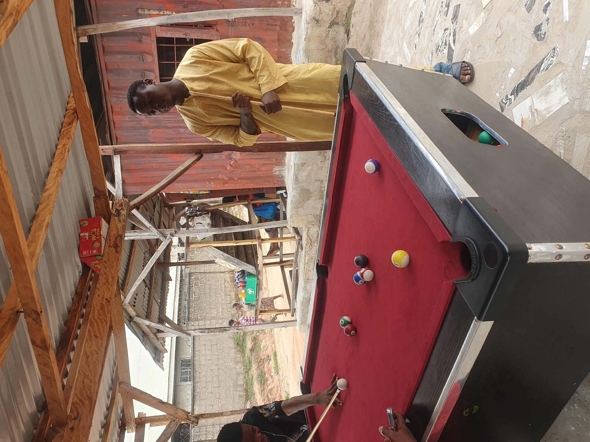 This is an outdoor image taken in Lokoja, Nigeria, depicting a casual game of pool. In the foreground, a red pool table with black trim is set up on a patterned tiled floor. Several pool balls are scattered on the table, including a white cue ball, a striped yellow ball, a solid yellow ball, a solid green ball, a solid red ball, and a solid blue ball with white stripes. A person in a black shirt is actively playing, in the process of striking the cue ball with a pool cue. To the right, a man wearing a long, yellow traditional garment stands holding a pool cue, observing the game. The background reveals a rustic outdoor setting with a corrugated metal roof supported by wooden beams, and partially constructed shelters. Further in the background, a red corrugated metal container structure is visible, along with a fence and some buildings. The overall environment suggests a local, informal gathering spot.