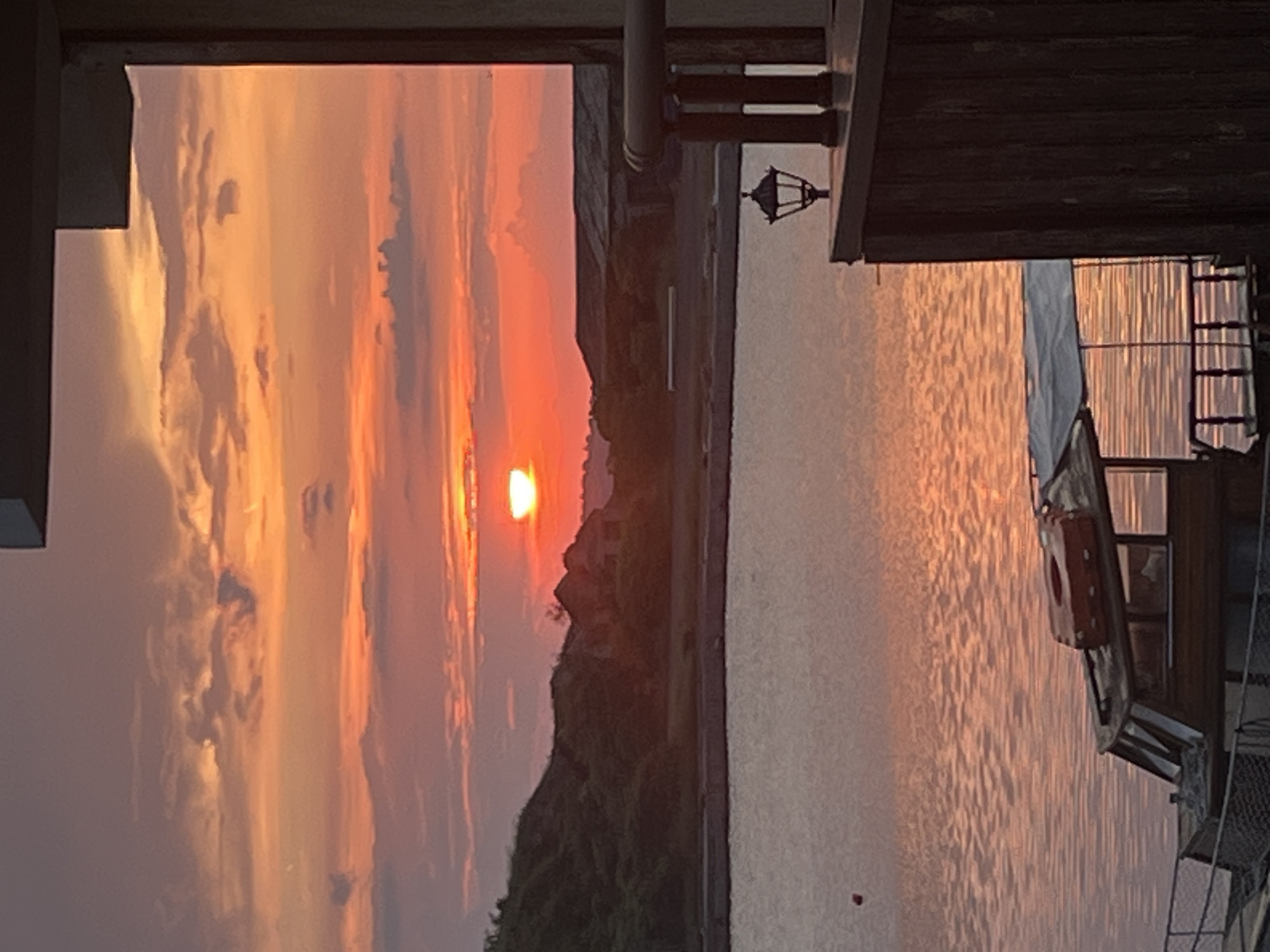 The image depicts a vibrant sunset over a body of water, likely a sea or large lake, viewed from what appears to be a pier or boat. The time of day is clearly sunset, with the sun's fiery glow dominating the sky.
In the sky, warm hues of orange, red, and yellow blend across the horizon, emanating from the partially visible sun on the left side of the frame. Scattered clouds, some dark and silhouetted, others illuminated by the sun, add texture to the colorful sky.
The expansive body of water fills the lower half of the image, reflecting the brilliant sunset colors in shimmering ripples across its surface. A dark, undulating silhouette of distant land or mountains stretches along the horizon, contrasting sharply with the bright sky above.
On the right side of the image, dark wooden structures are visible, suggesting a boat's cabin, a pier building, or a dock. A traditional-style lamppost with an ornate lantern stands on this structure. An orange and white lifebuoy is attached to the side of the wooden structure, and glimpses of railings or ladders can be seen further to the right. The upper left corner shows a dark, angular architectural element, possibly an overhang, framing the scene.
The overall atmosphere is serene and picturesque, highlighting the beauty of nature at dusk. There is no visible text in the image.