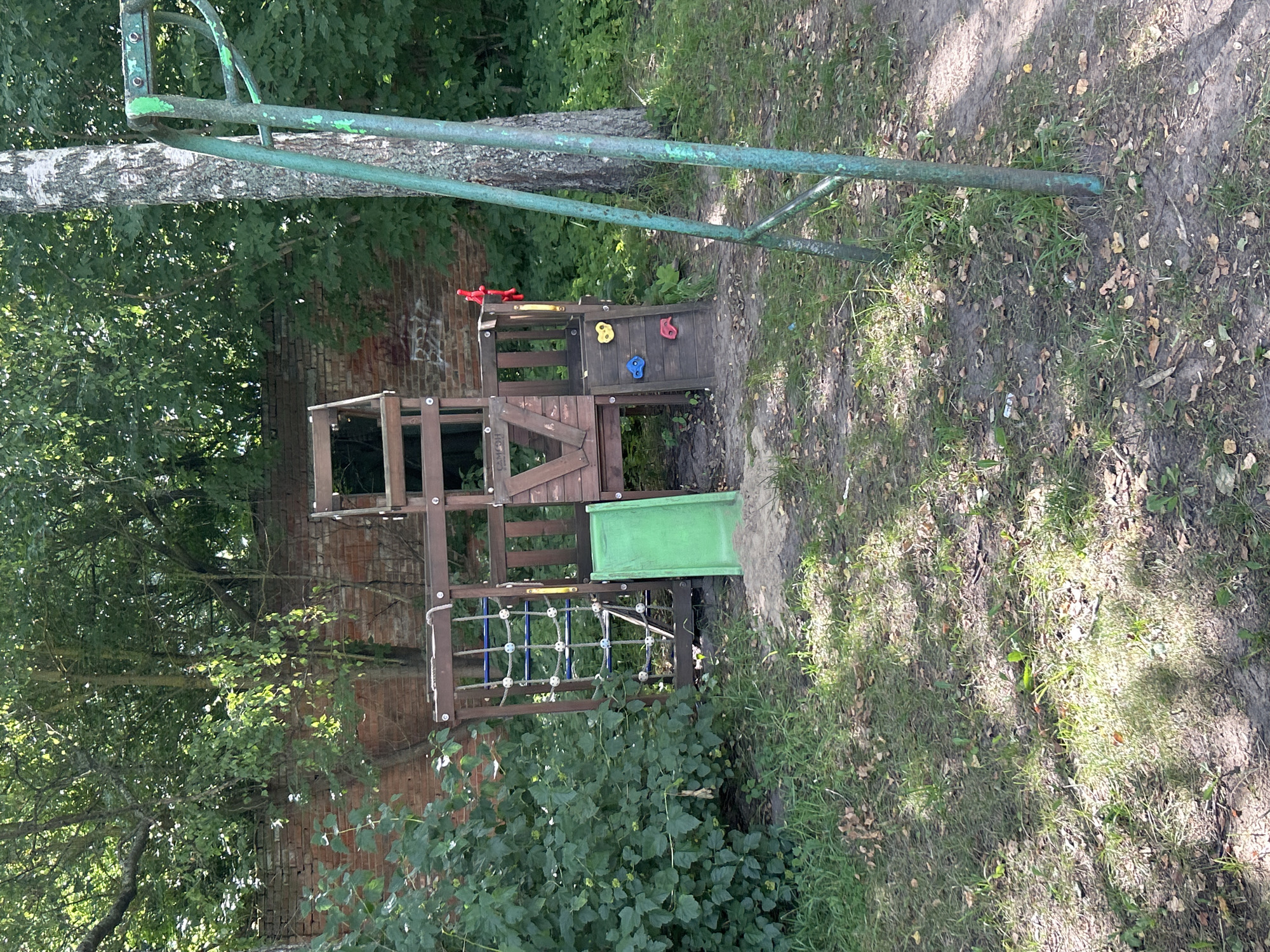 The image shows an outdoor playground area. In the center, a wooden multi-level play structure is visible. It features a rope net climbing wall on the left, a green slide on the front, and climbing holds on the right. A portion of a green metal swing set frame is present in the foreground on the right. The playground is situated in a grassy area with scattered leaves and dirt. Behind the play structure, a brick wall and dense green foliage are present, with a birch tree trunk to the right of the play structure. The overall scene suggests a children's play area within a naturalistic setting, possibly in a park or backyard. The location is identified as Kalitsino, Russia.