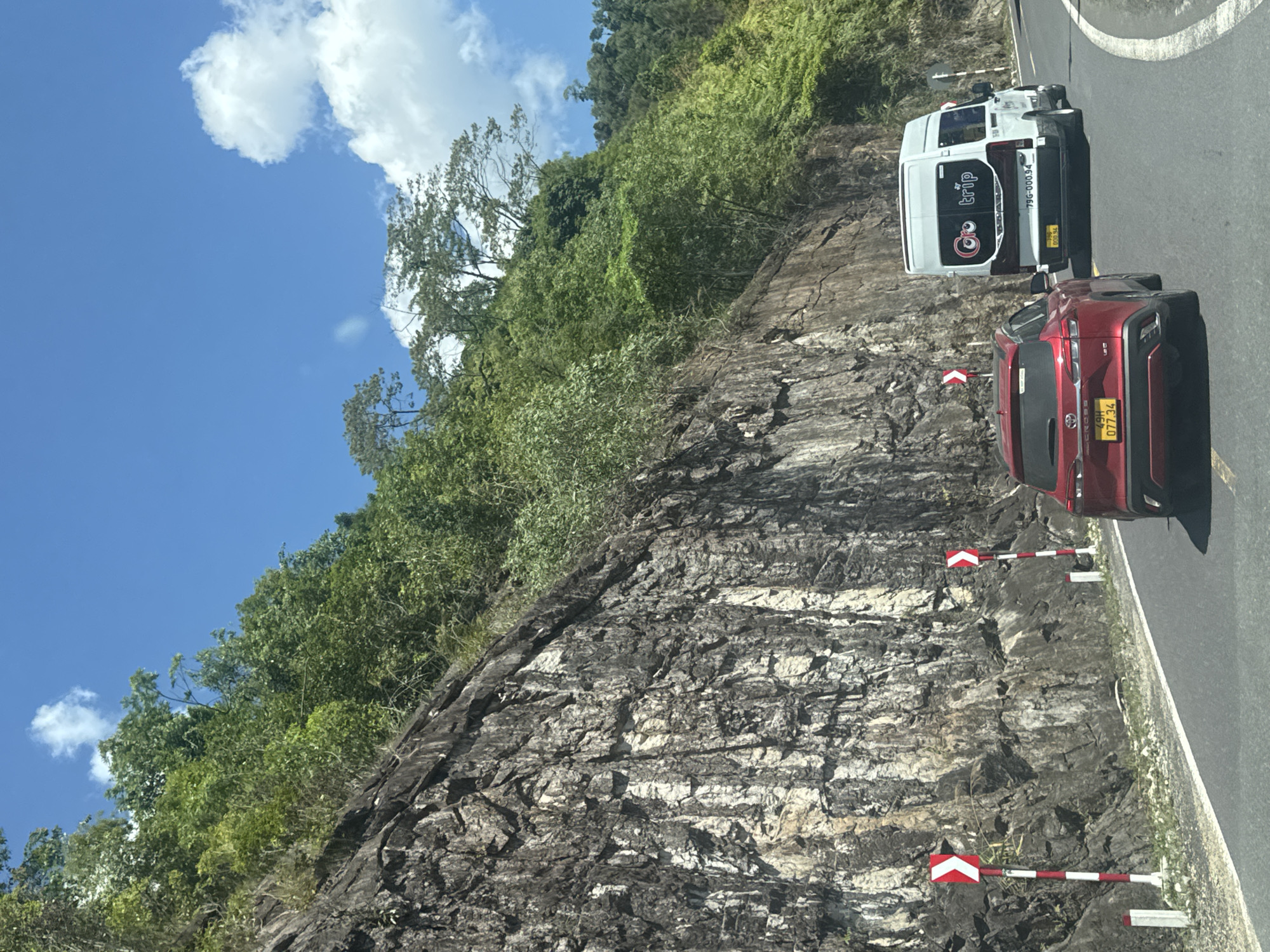This is a medium-shot, eye-level, outdoor, daytime, vertical photograph taken from a vehicle moving on a paved road. The road curves to the right and is bordered by a steep, rocky cliff face on the left and a dense, green hillside on the right. The cliff face is composed of rough, grey rock with visible cracks and striations, interspersed with patches of lighter colored minerals. Green foliage, including trees and bushes, grows from the top and crevices of the cliff. The hillside to the right is densely vegetated with various shades of green trees and plants.
Two vehicles are visible on the road. In the foreground, a red SUV is moving away from the camera, angled slightly to the right. It has a license plate that reads "49H 077 34". To the right of the SUV, a white passenger van is also moving away from the camera. It has "GoTrip" written in a circular logo on its rear window and a license plate that reads "79G-000.94".
Roadside markers with red and white stripes and a red chevron are present on the left side of the road, indicating a curve. The sky is a vibrant blue with scattered white clouds. The scene appears to be a scenic mountainous or hilly area, possibly a mountain pass. The general location is identified as Nam Khanh Vinh, Vietnam. No people are visible.