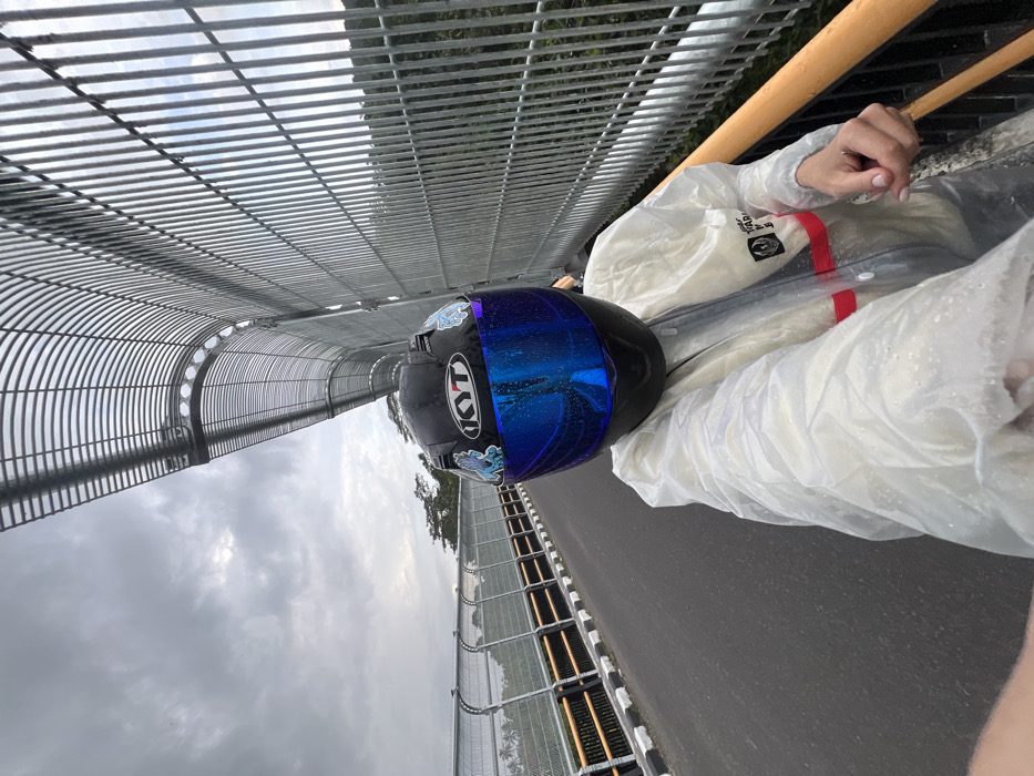 A person is depicted in a close-up, self-portrait perspective on a bridge located in Sembung, Indonesia. The individual wears a black KYT full-face helmet with a reflective blue visor, adorned with light blue cartoon-like stickers. A translucent white raincoat, covered in small water droplets indicating recent precipitation, is worn over other clothing, displaying a partial logo and a red horizontal stripe on the chest. The person's left hand, with a ring on the ring finger and light-painted nails, is visible in the lower-left foreground, holding the device used to capture the image.
The environment consists of an asphalt bridge surface. To the left, a black and white striped curb borders the road, followed by a guardrail with orange-brown horizontal elements, silver vertical posts, and a wire mesh fence. To the right, a prominent, tall metal barrier composed of vertical bars and an arched overhead grating creates a partial tunnel-like enclosure along the bridge's edge. Beyond the bridge's structures, green trees and lush foliage are visible, suggesting a natural landscape. The sky is overcast with grey clouds.