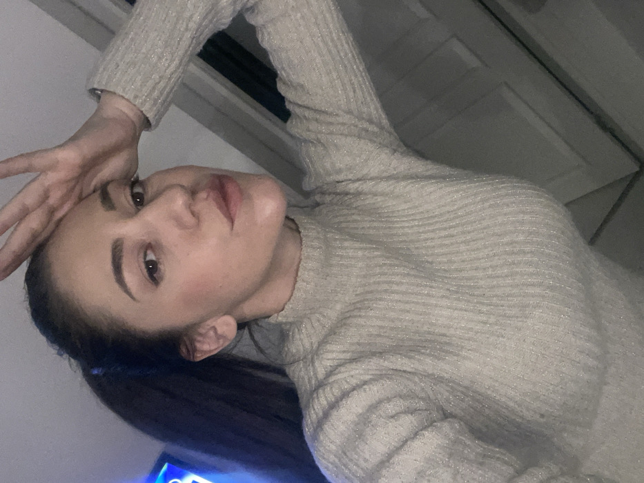 An adult female is depicted in a close-up, indoor setting. She has dark hair pulled back into a high ponytail, visible on the left side of the frame. She is wearing a light-colored, ribbed turtleneck sweater, possibly beige or pale grey, which exhibits a subtle metallic sheen. Her right hand is raised to her forehead, fingers slightly splayed, with the thumb near her temple. Her gaze is directed towards the camera. The background is dimly lit and blurred, featuring a light-colored, paneled surface, possibly a door or wall, on the right. A diffused, blue light source or object is partially visible in the upper left background. No specific visual markers or landmarks are present within the image to identify a precise location within the city of Sofia, Bulgaria.