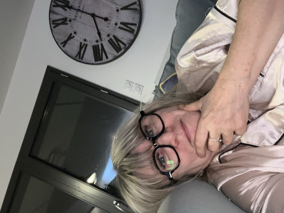 A woman wearing glasses and a light pink satin pajama set is lying on a blue pillow. Her hand is near her face. Behind her is a closed black-framed glass door. Above the door hangs a round, dark-rimmed clock with Roman numerals. The clock's hands show approximately 10:25 or 2:55. Two white electrical outlets are positioned on the wall above the door frame.