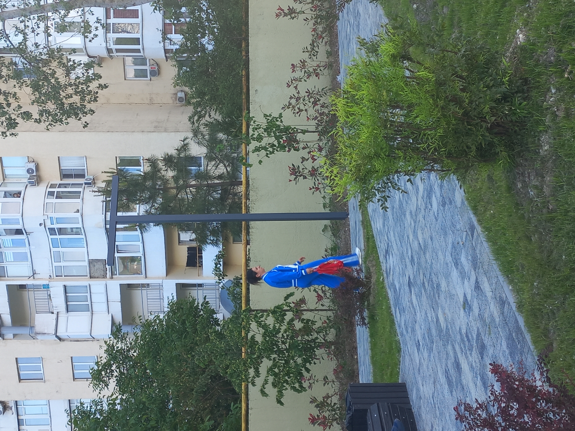 A medium shot of a paved walkway in the city of Unknown, Unknown, with a person dressed in a blue tracksuit standing on the walkway. The person is holding red objects and is positioned between trees and bushes, with a tall building in the background. The sky is not visible in the image.