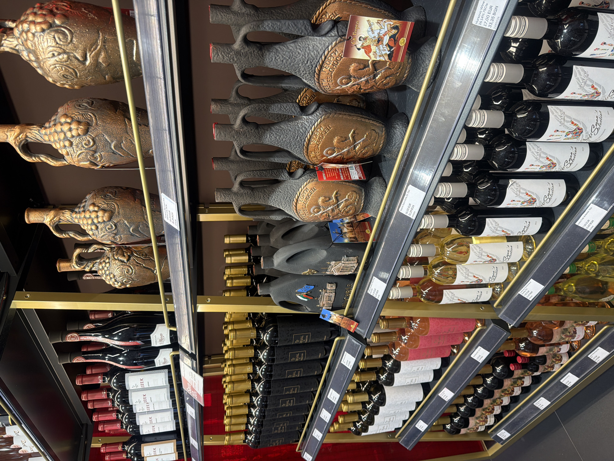 This is a medium shot, eye-level, indoor, eye-level photograph of a retail display of alcoholic beverages, primarily wine, in a store located in Sofia, Bulgaria. The display consists of multiple shelves filled with various types of wine bottles, some with dark glass and red necks, others with lighter glass and gold or silver caps. In addition to bottles, there are decorative ceramic jugs with intricate designs, some appearing to be dark grey and others bronze-colored. Some jugs are adorned with decorative motifs and even small flags.
Price tags are visible on several shelves, indicating prices in EUR and BGN, confirming the location as Bulgaria. One prominent price tag reads "Mavro Sant Sofia 75cl 16,00 EUR 31,29 BGN". Another reads "Hvrosovo wine 26,14% 75cl 17,00 EUR 33,25 BGN".
The lighting is artificial and appears to be from overhead store lights, creating some reflections on the bottles and jugs. The background is a dark red wall with metallic gold shelving units supporting the displays. The overall presentation suggests a wine shop or a section of a larger supermarket. No people are visible in the image.