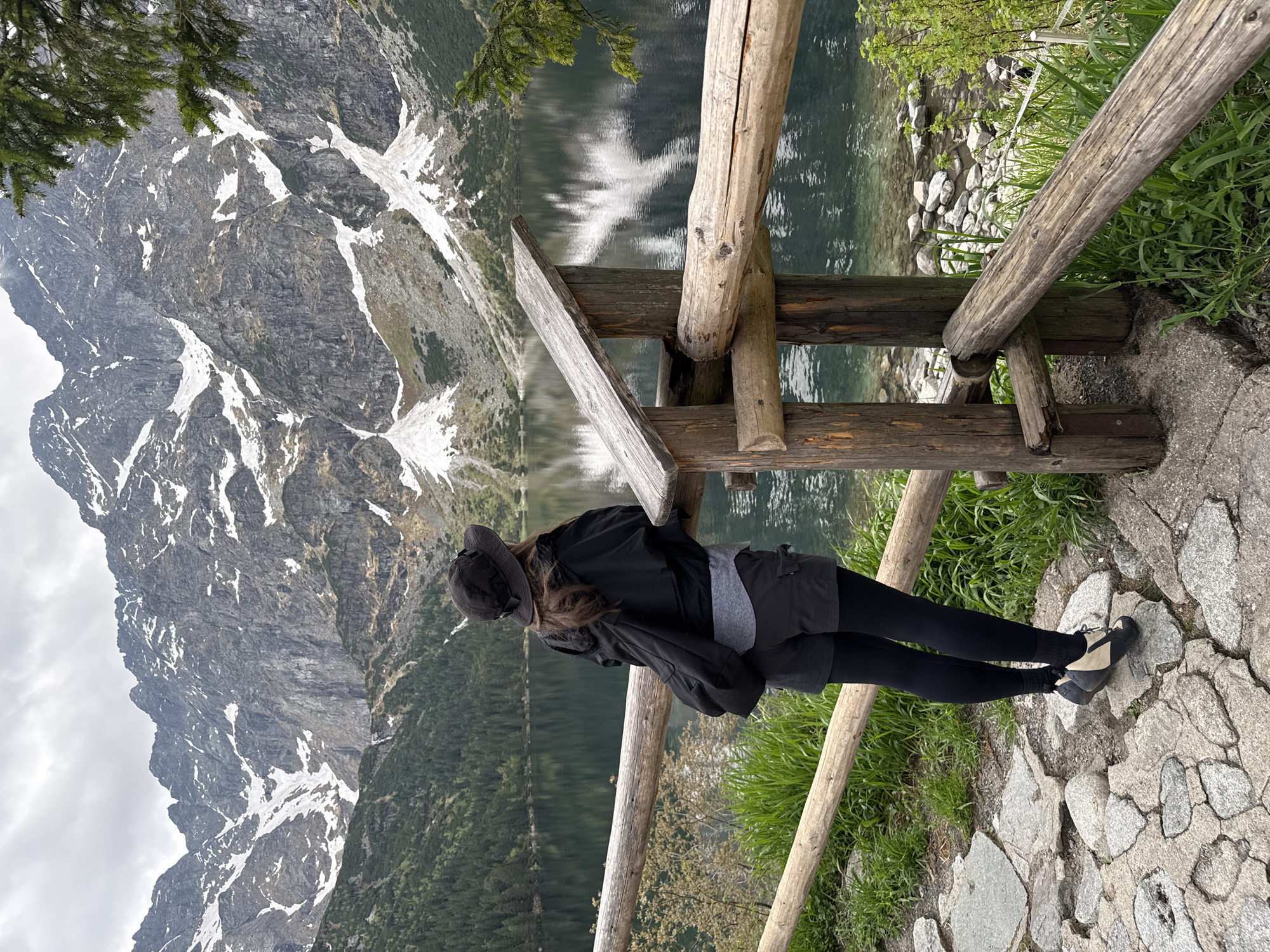 A woman in black leggings, grey shorts, a grey shirt, and a black jacket leans on a wooden fence overlooking a lake reflecting snowy mountains. The scene is in Brzegi, Poland. The ground is a stone path. The weather appears overcast.